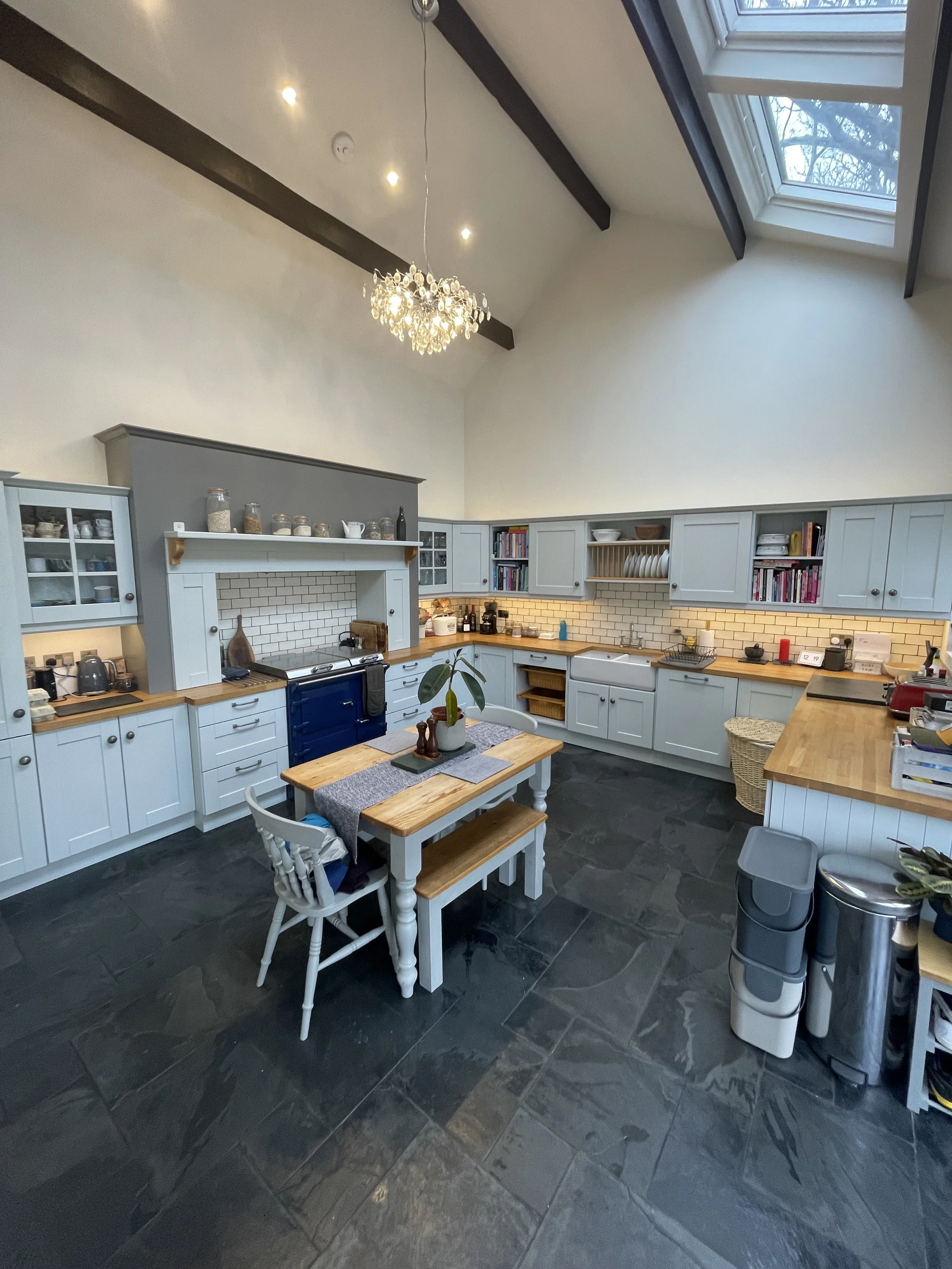 A spacious kitchen with light blue cabinets, wooden countertops, and a dark slate floor. There is a small dining table with chairs and a bench, a chandelier, and a ceiling with skylights.