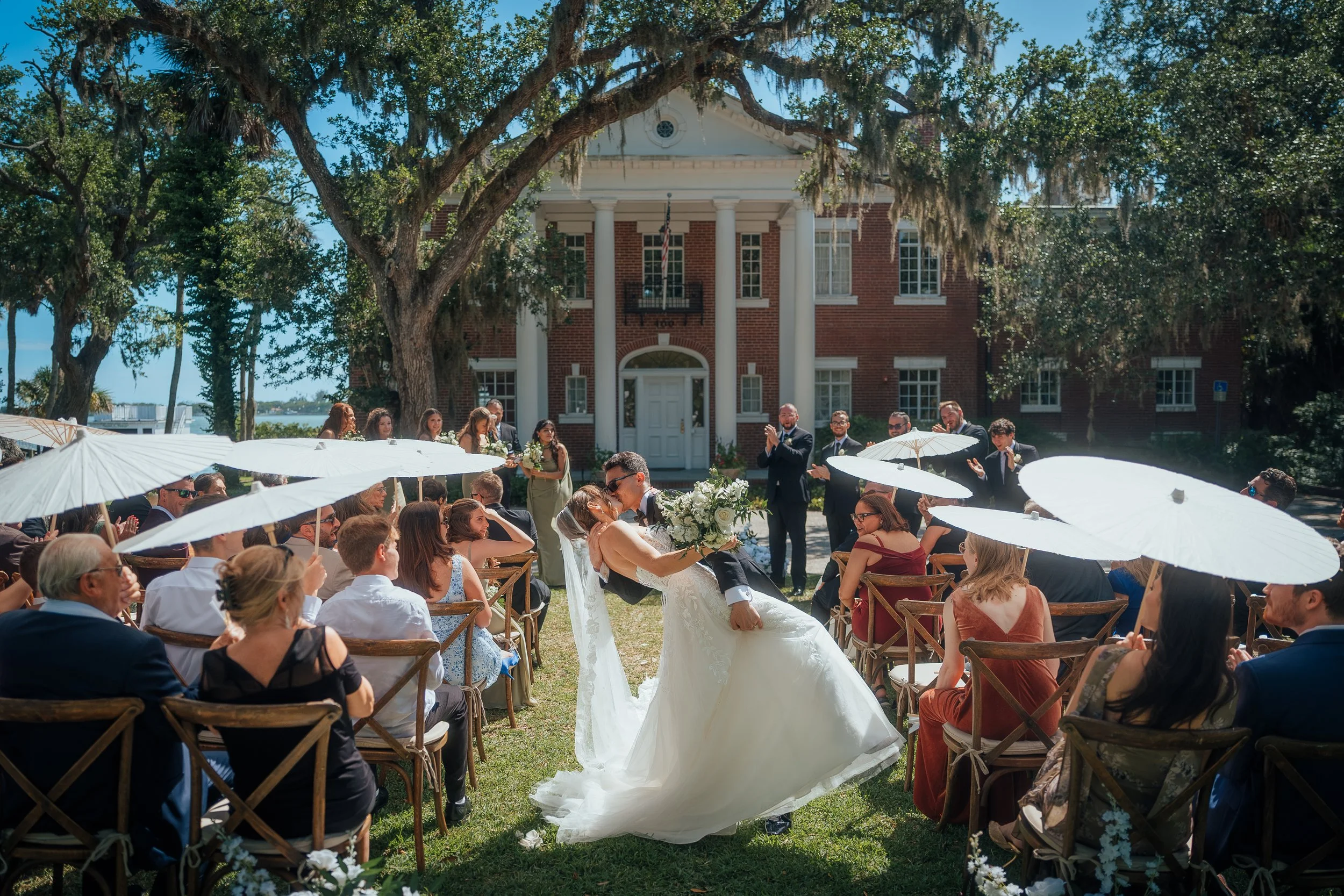 A wedding ceremony outdoors at the Bay Preserve, a historic venue. The bride and groom are kissing while guests sit under white umbrellas and watch.