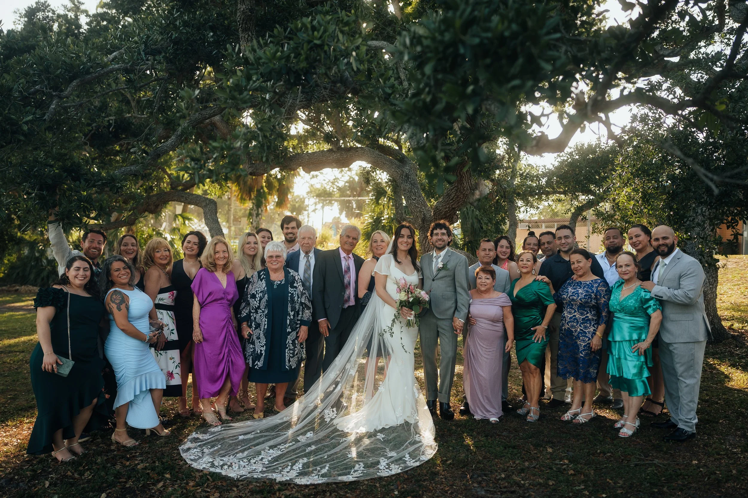 A large, diverse group of people gathered outdoors under a big tree for a wedding celebration, with the bride and groom in the center. The bride is wearing a white wedding dress with a long train, holding a bouquet, and the groom is dressed in a suit. The group is smiling and posing for a photo during the golden hour.