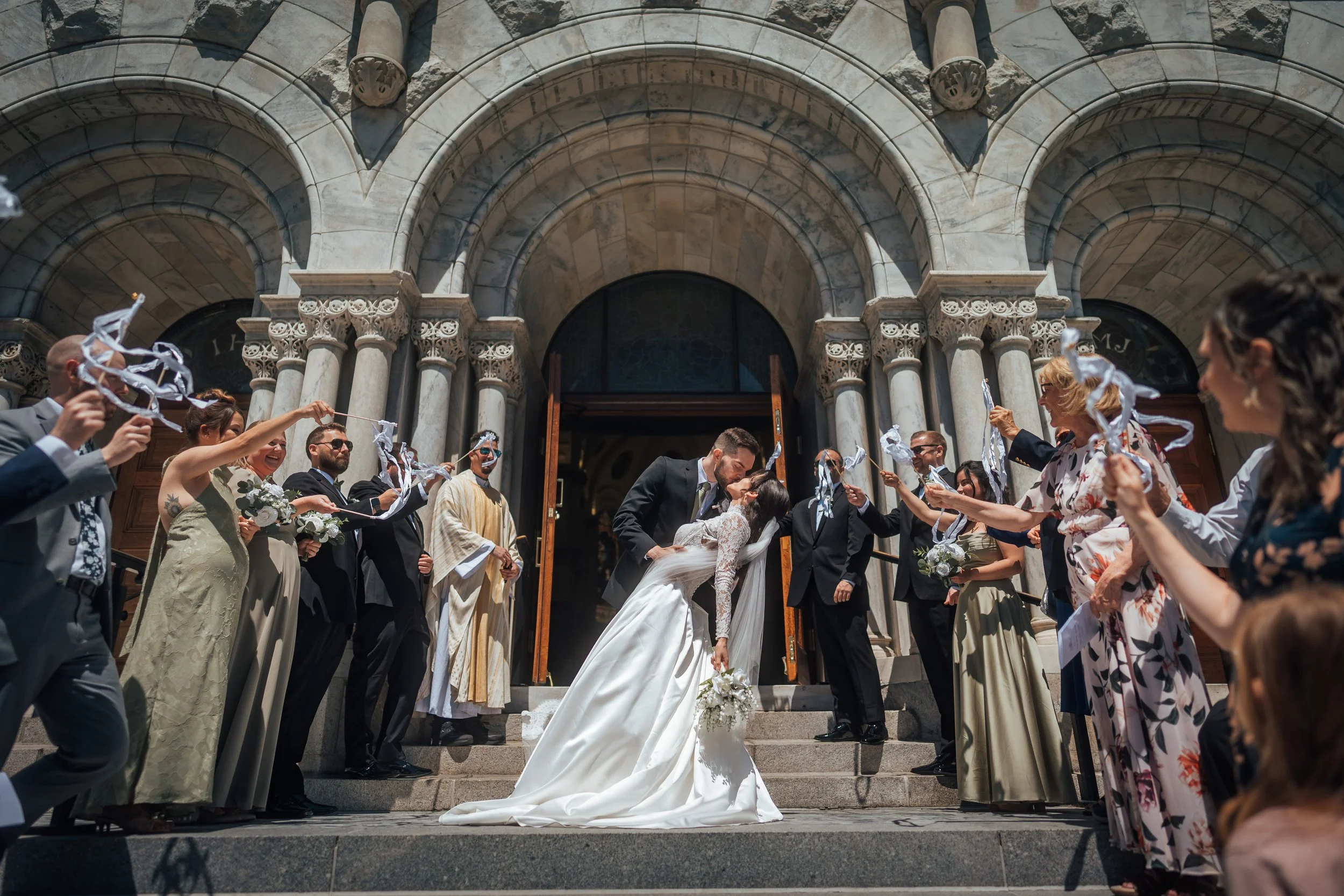 A wedding ceremony taking place on the steps of Sacred Heart Catholic Church in Tampa. The bride and groom are kissing while surrounded by guests, some holding paper streamers. The scene is festive and sunny.