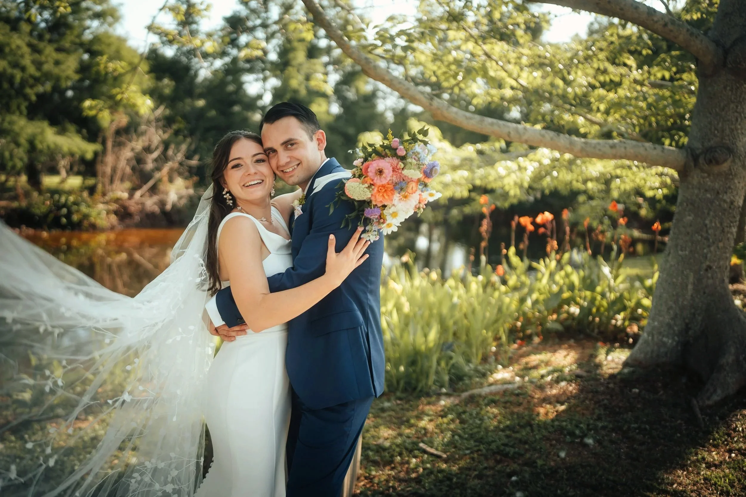 A bride and groom smiling and embracing outdoors at Palma Sola Botanic Park under a tree, with the bride holding a colorful bouquet of flowers, during their wedding celebration.
