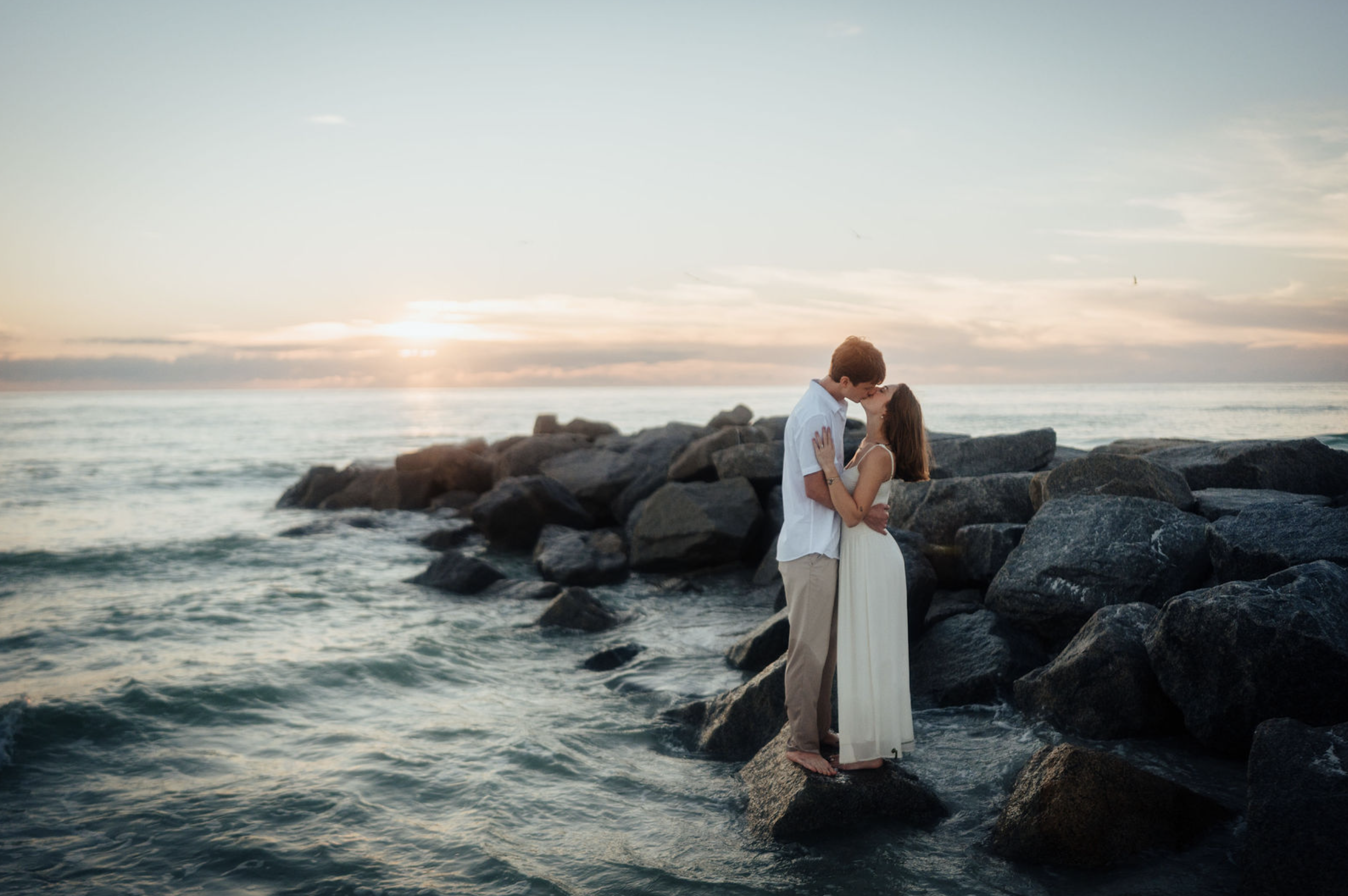 A couple standing barefoot on rocks at south Lido Key beach, by the ocean at sunset, embracing and about to kiss.