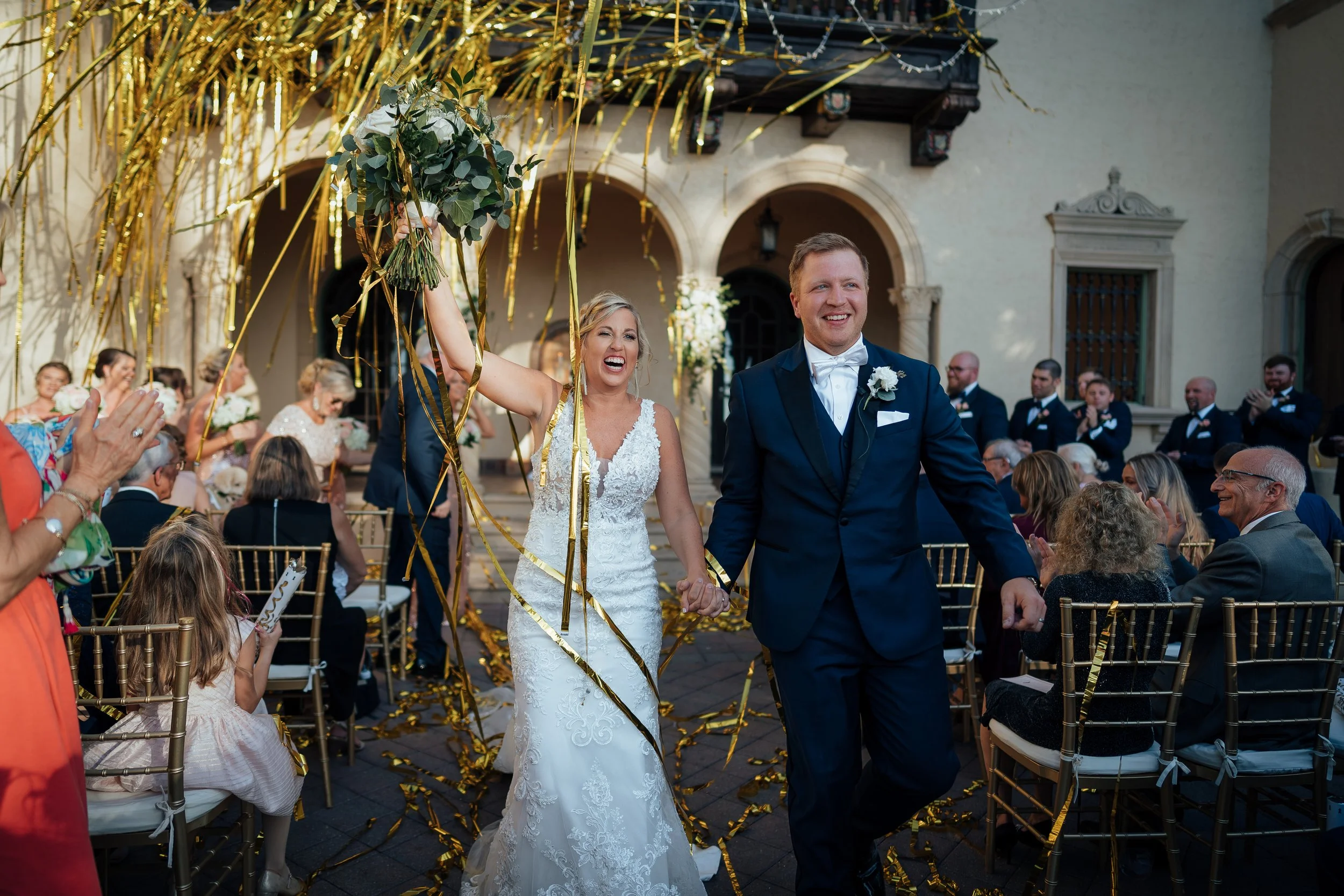 A newlywed couple walks hand-in-hand at their wedding ceremony in the courtyard of the Powel Crosley in Sarasota, Florida, with guests seated around. Gold streamers and confetti decorate the scene, with guests clapping and celebrating.