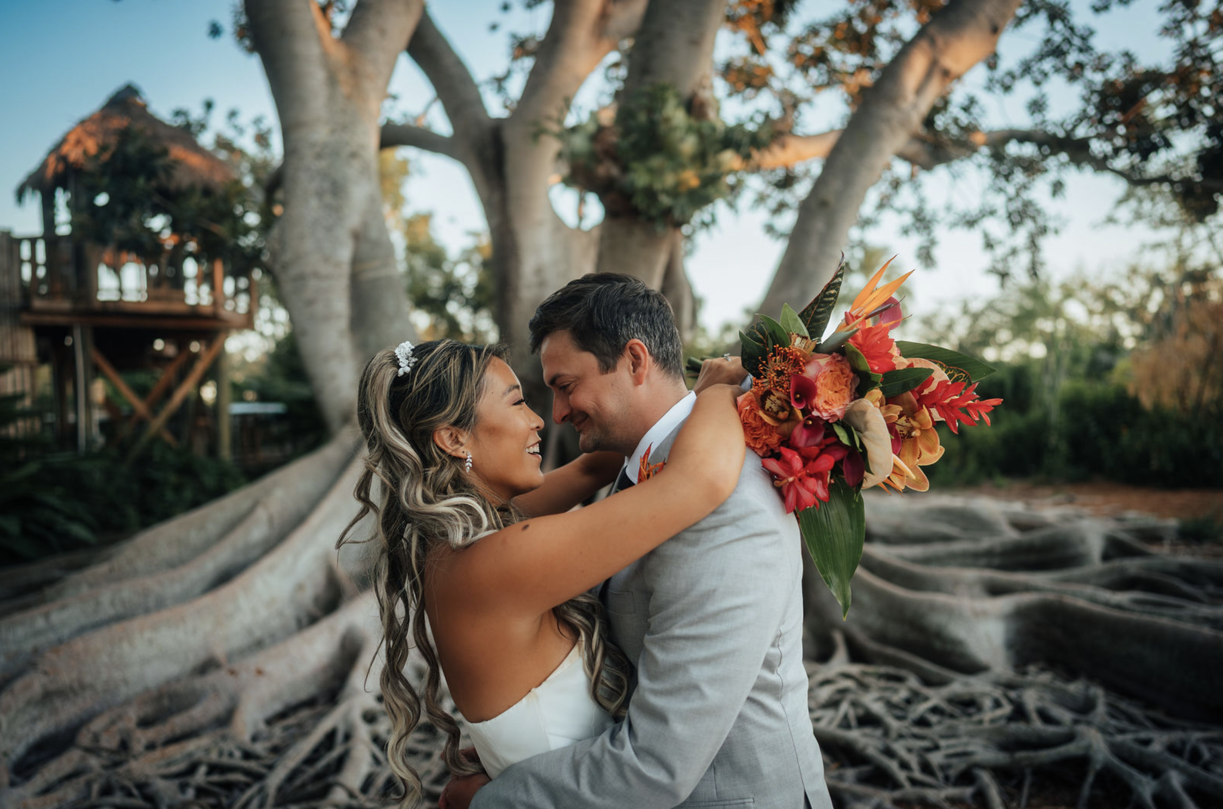 Candid wedding portrait at Selby Gardens. A newlywed couple embraces under a large banyan with the bride holding a bouquet of vibrant orange, red, and pink flowers. 