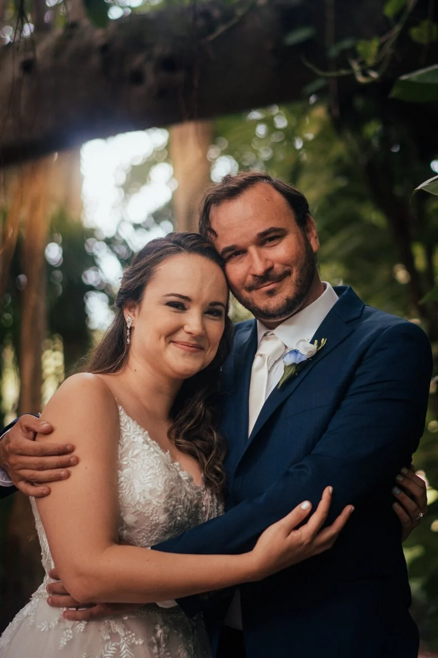 A happy bride and groom embracing outdoors surrounded by greenery, smiling at the camera.