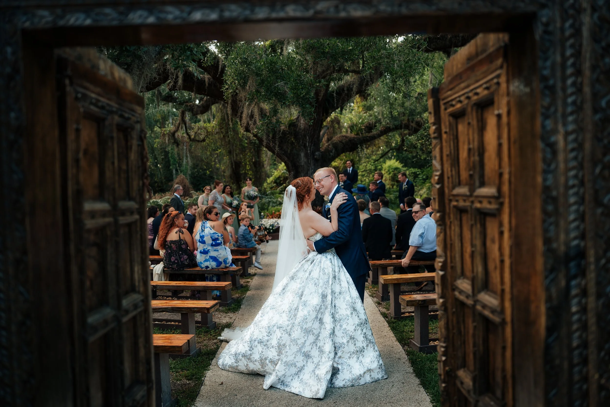 A wedding ceremony taking place outdoors at Del Favero Oasis in Tampa, Florida framed by a decorative wooden gate, with a bride and groom embracing in front of a large tree and seated guests in the background.