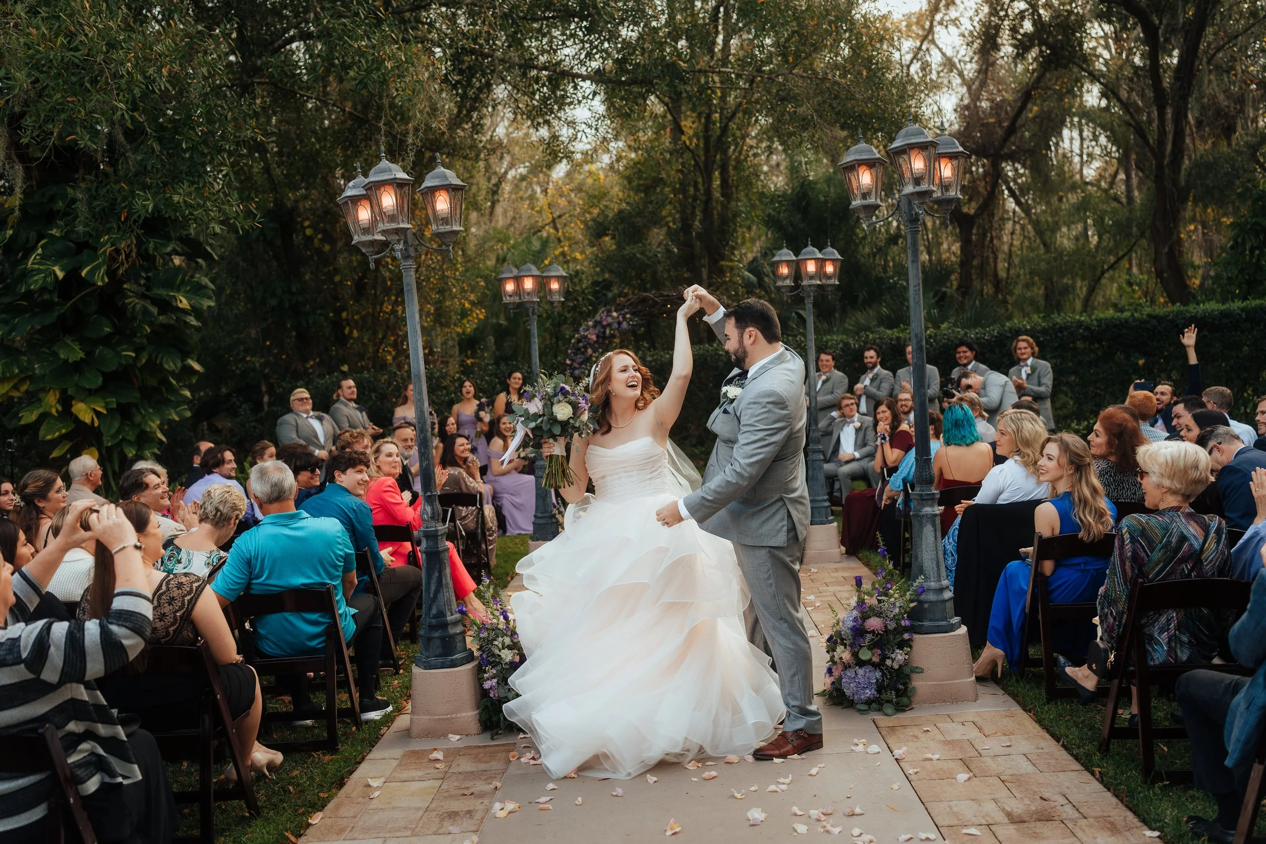 A bride and groom dancing under street lamps at their outdoor Bakers Ranch wedding ceremony, surrounded by seated guests and lush greenery.