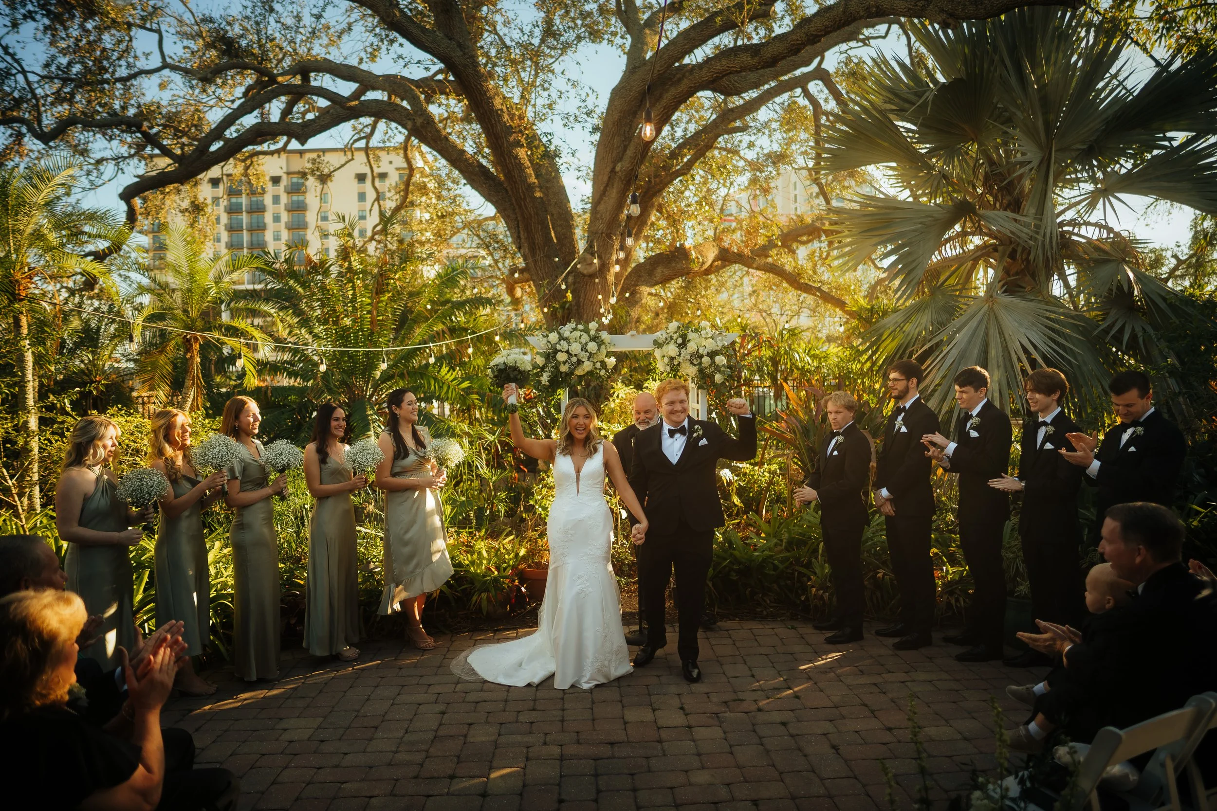 A bride and groom holding hands and celebrating during their wedding ceremony outdoors at the Sarasota Garden Club in a lush garden with string lights and tall trees.