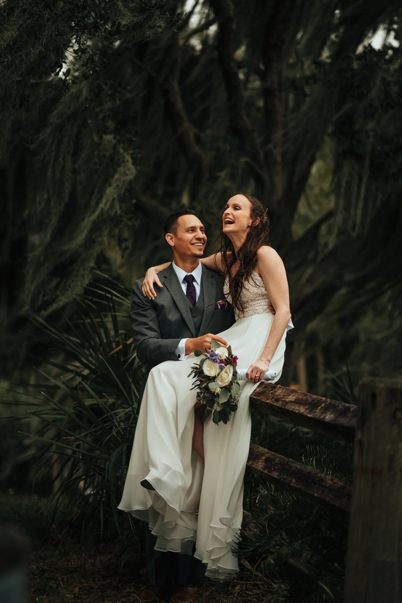 Bride holding a bouquet smiles beside the groom on a log at Royal Pine Estate in Seminole, Florida.