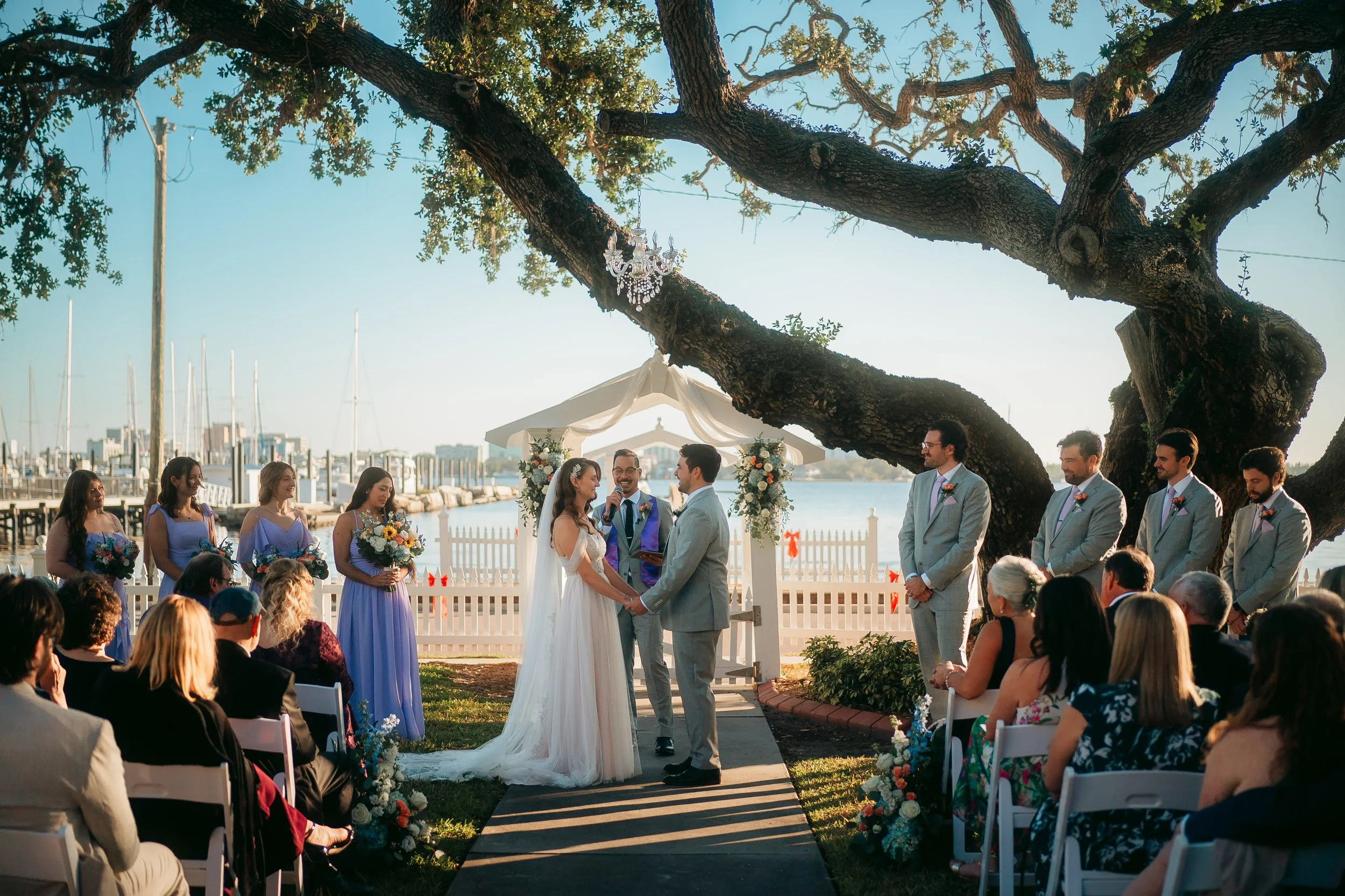 A wedding ceremony outdoors at the Palmetto Riverside Bed and Breakfast, by the water under a large tree with a crystal chandelier hanging from its branches. The bride and groom are exchanging vows.