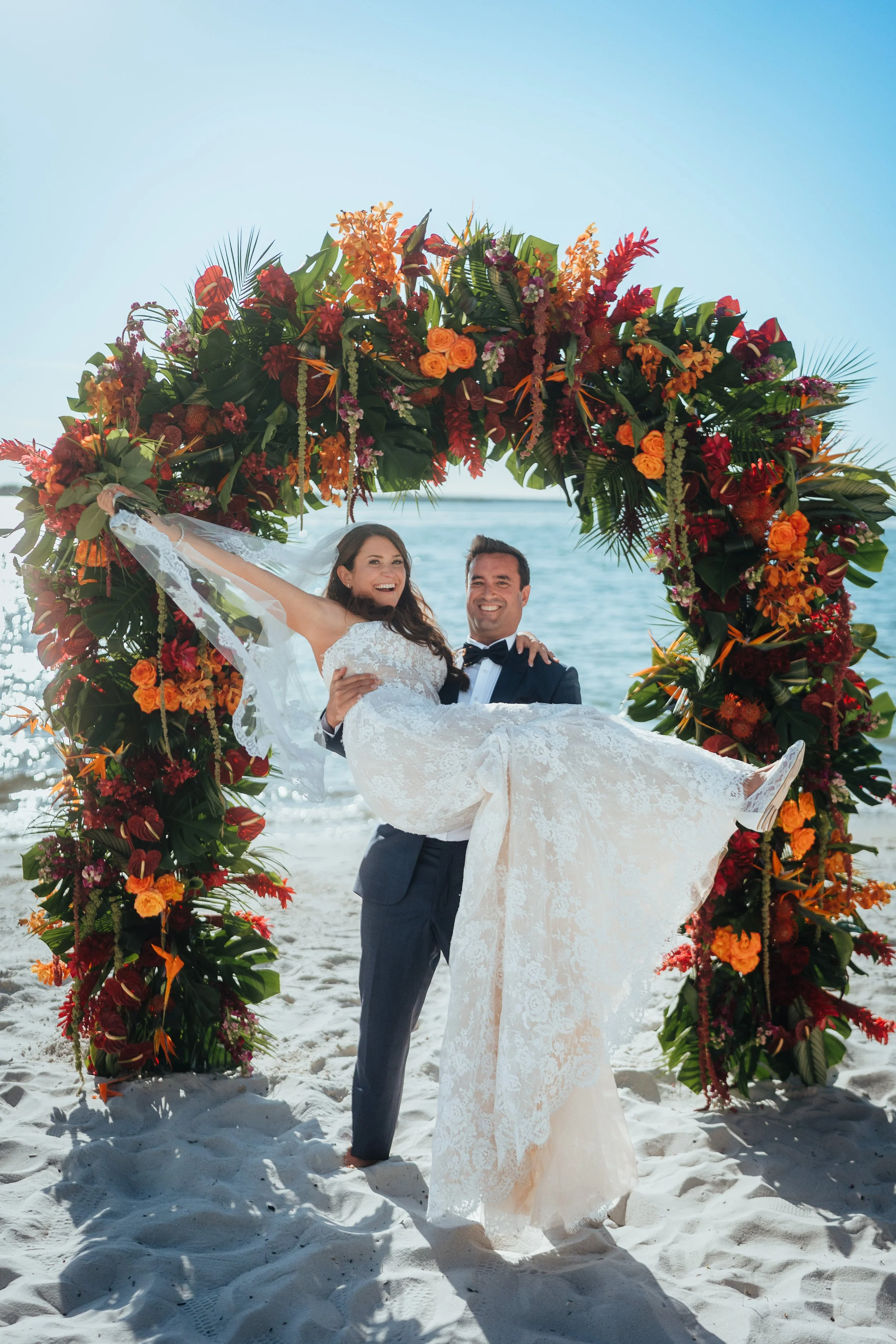 A newlywed couple at Sannibel beach, the groom in a dark suit and the bride in a white lace wedding dress, standing under a large floral arch with orange and red flowers, with the ocean and clear sky in the background.