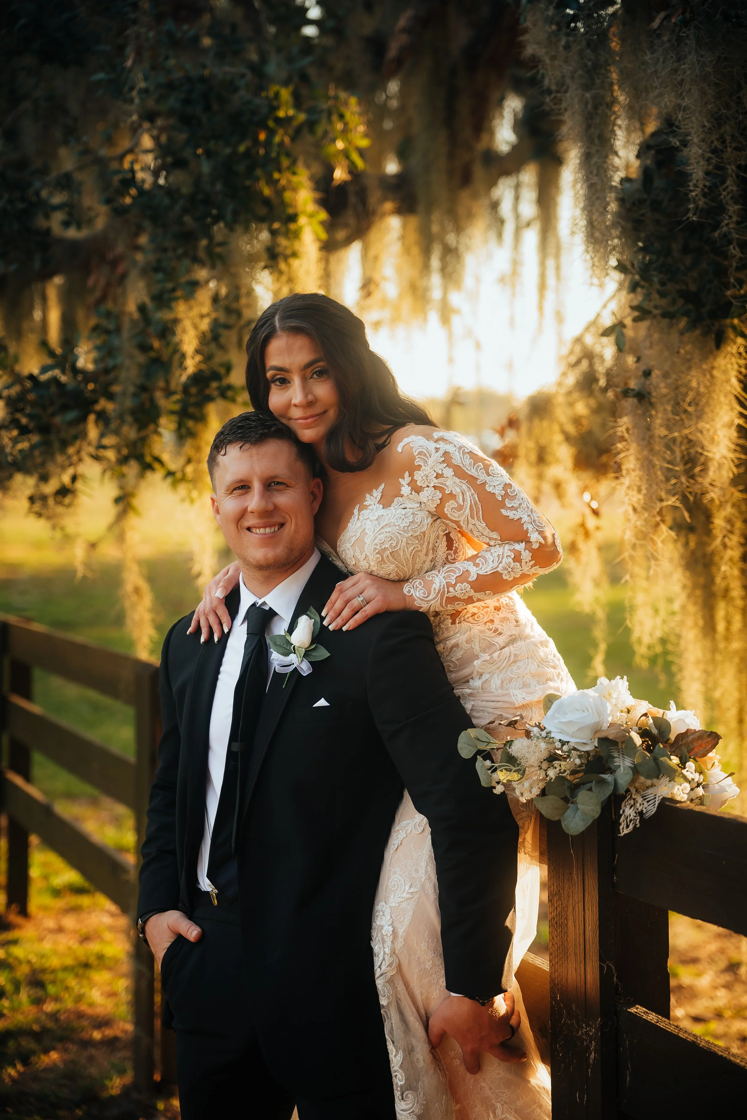 A bride and groom pose outdoors during sunset on the grounds of Bella Cosa wedding venue, surrounded by hanging moss and trees, with the bride wearing a lace wedding dress and the groom in a black suit and tie, both smiling.