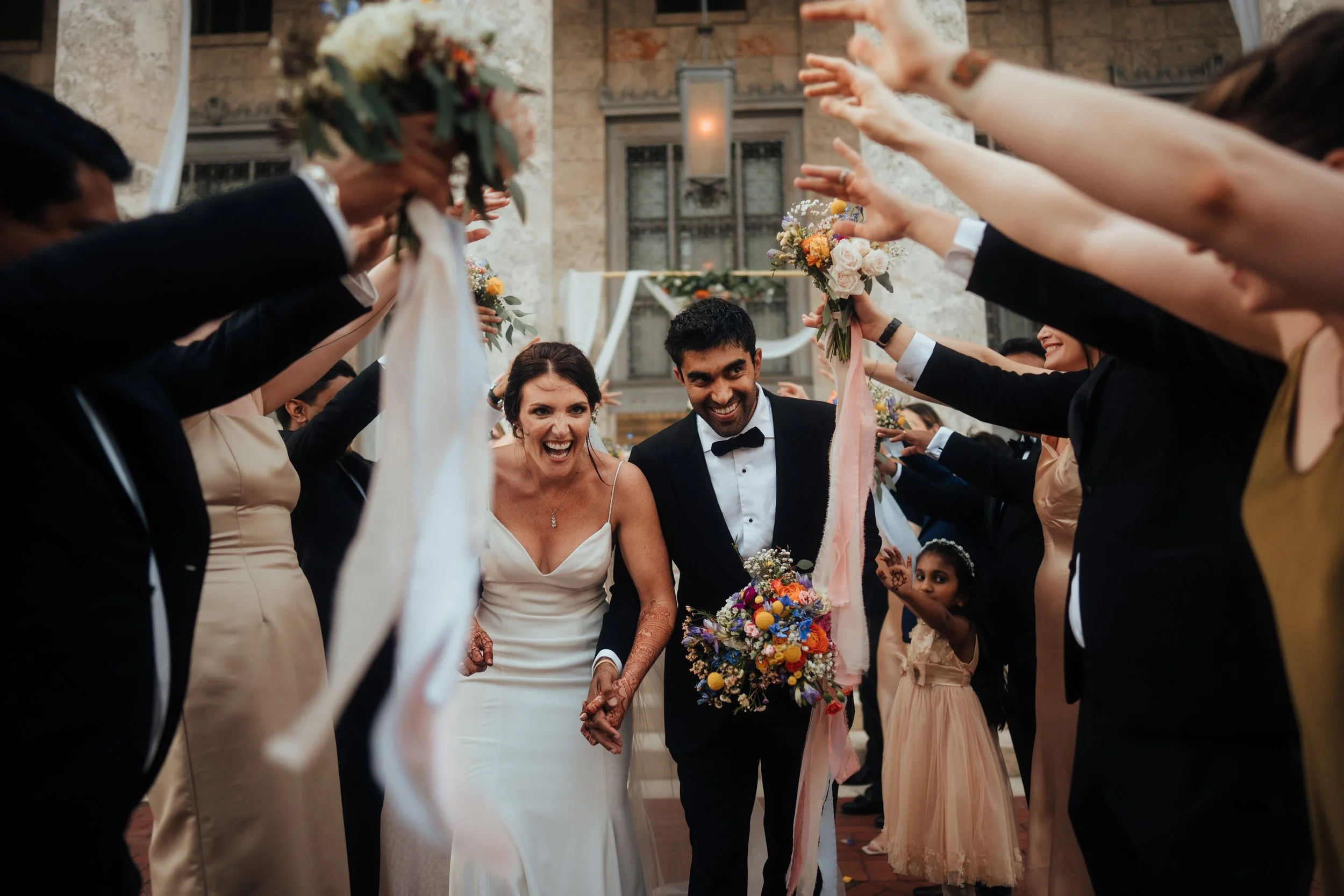 A bride and groom holding hands and smiling as they walk down the steps of the Sidney & Berne Davis Art Center through a tunnel of bridesmaids and groomsmen during their wedding celebration inside a historic building.