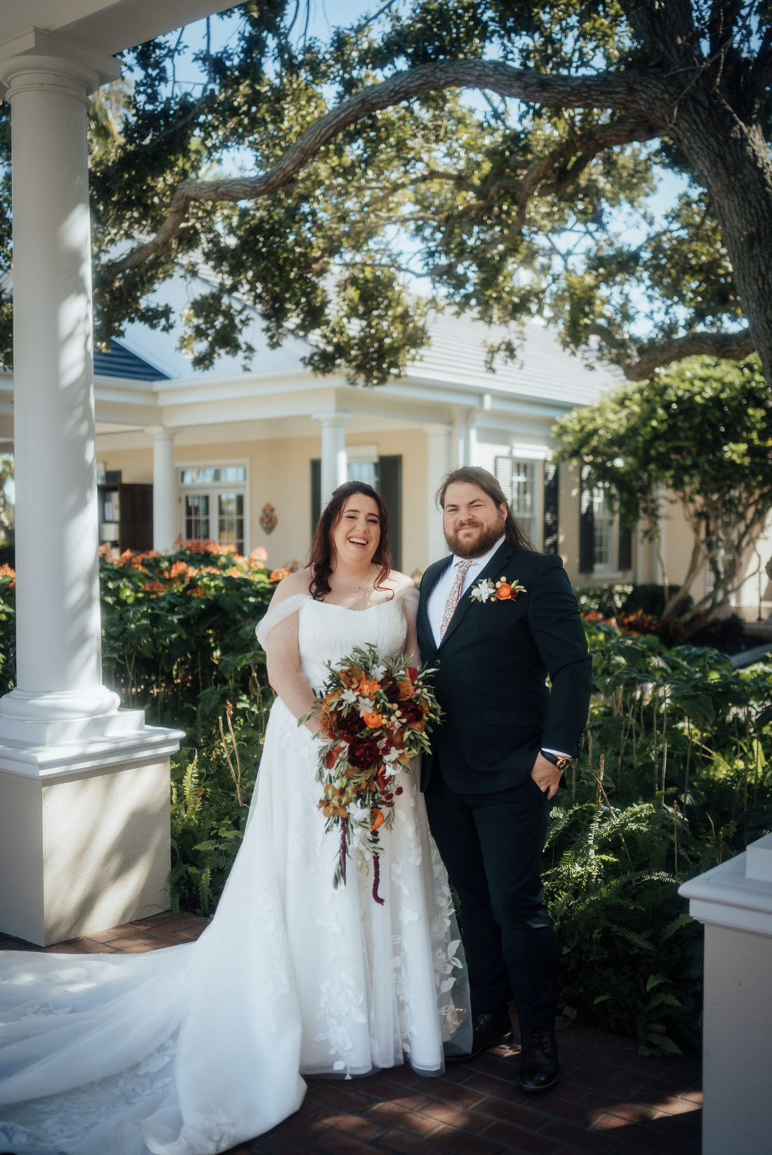 A newlywed couple stands outside in front of The Oaks Country Club in Osprey.  The woman wears a white wedding gown and holds a cascading bouquet of red, orange, and white flowers. The man wears a black suit with a floral boutonniere. 