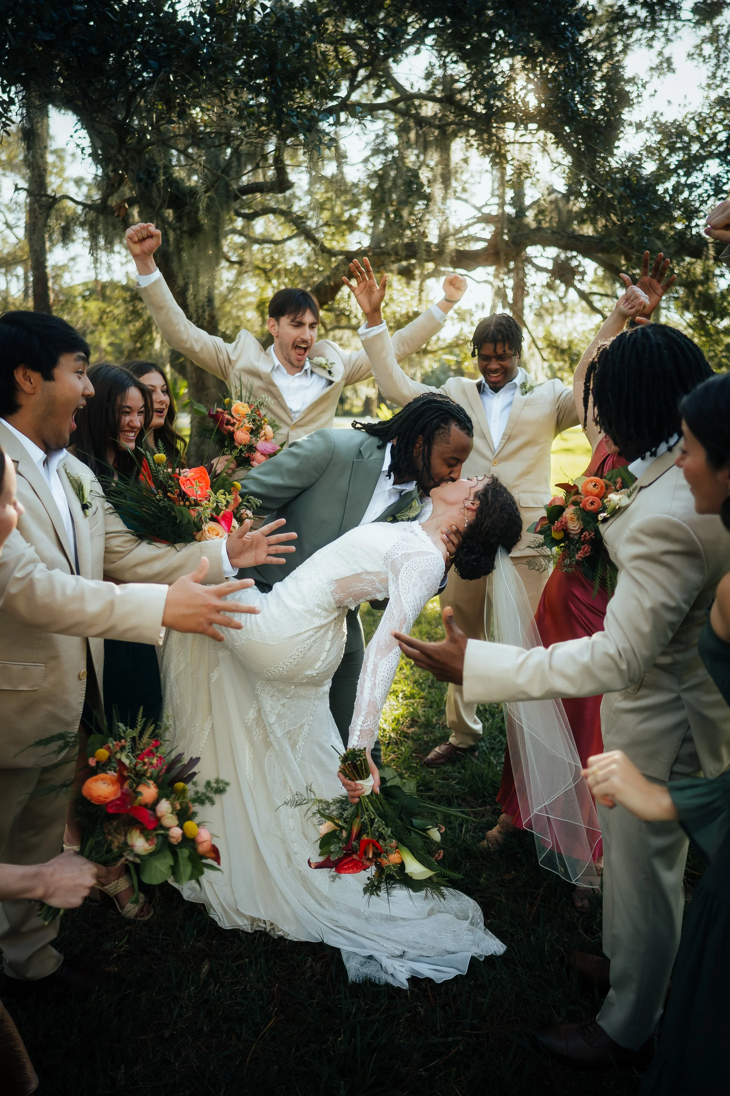 An intimate wedding celebration outdoors with a bride and groom kissing, surrounded by friends and family holding bouquets, under large trees and sunlight at Royal Pine Estate in Tampa, Florida.