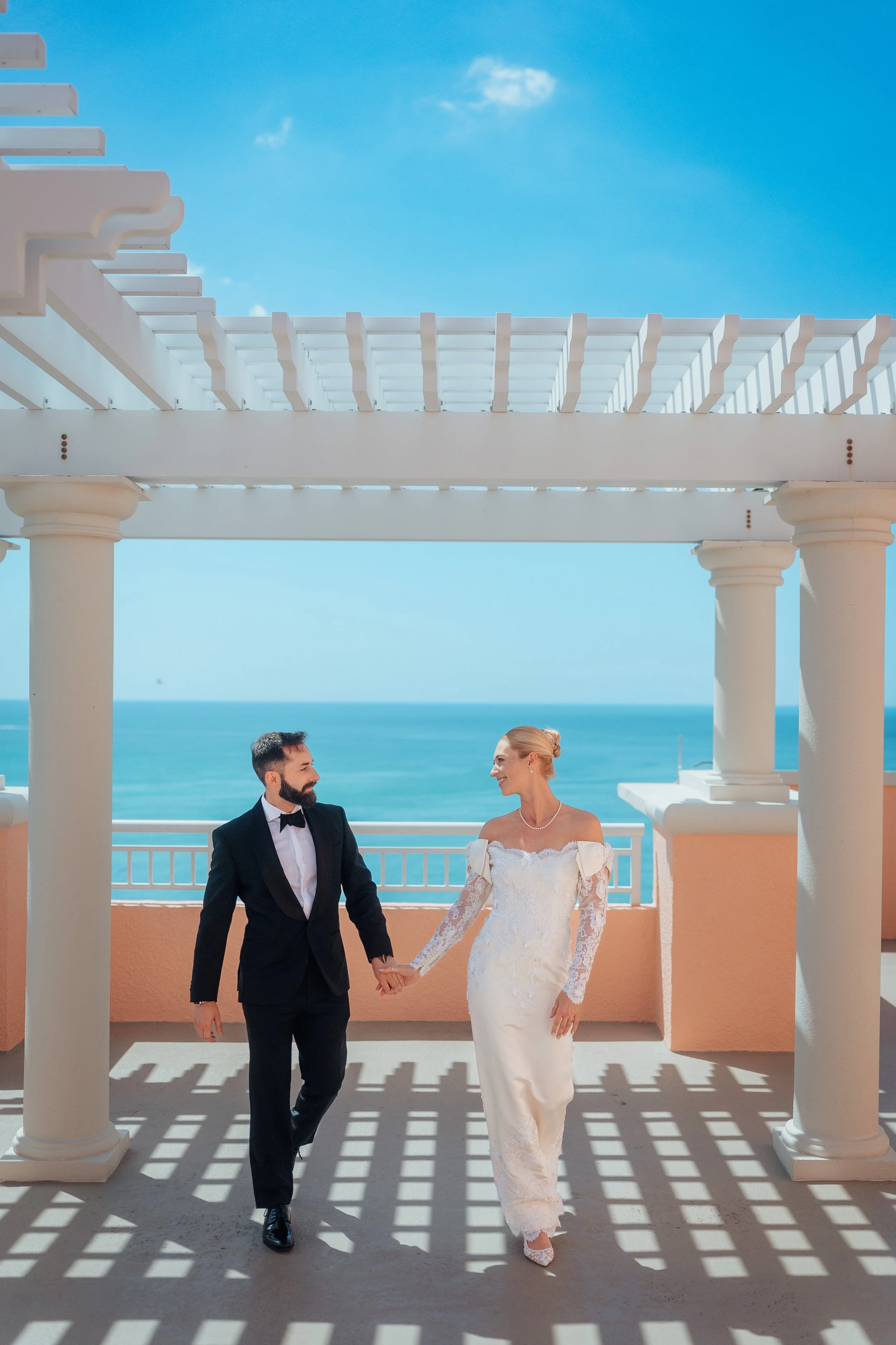 A bride and groom holding hands while walking under a white pergola on the balcony of the Hyatt Clearwater by the seaside on a sunny day.