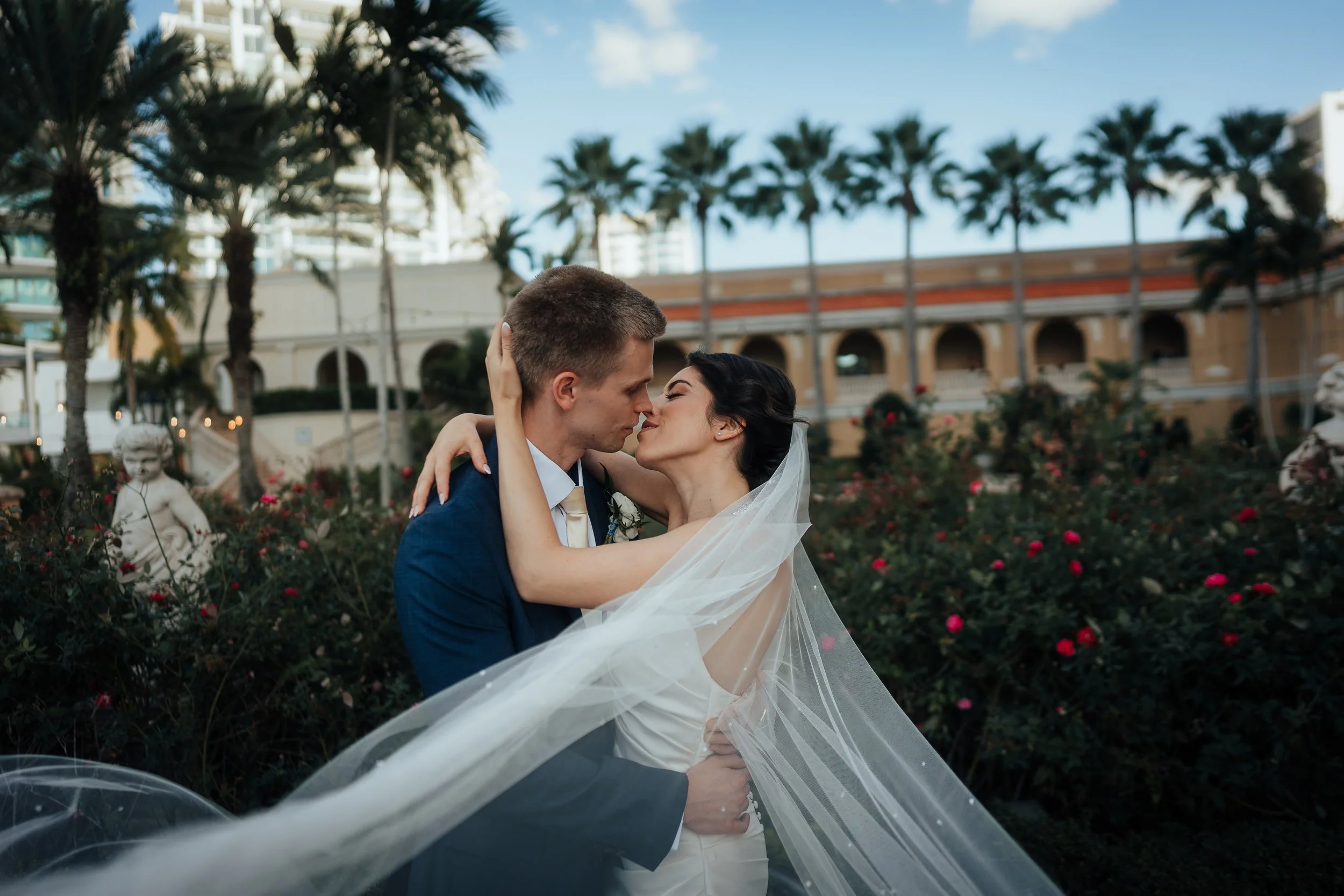 Ritz Carlton Sarasota wedding portrait. A bride and groom are sharing a romantic moment outdoors, surrounded by flowers, palm trees, and statues with a building in the background.