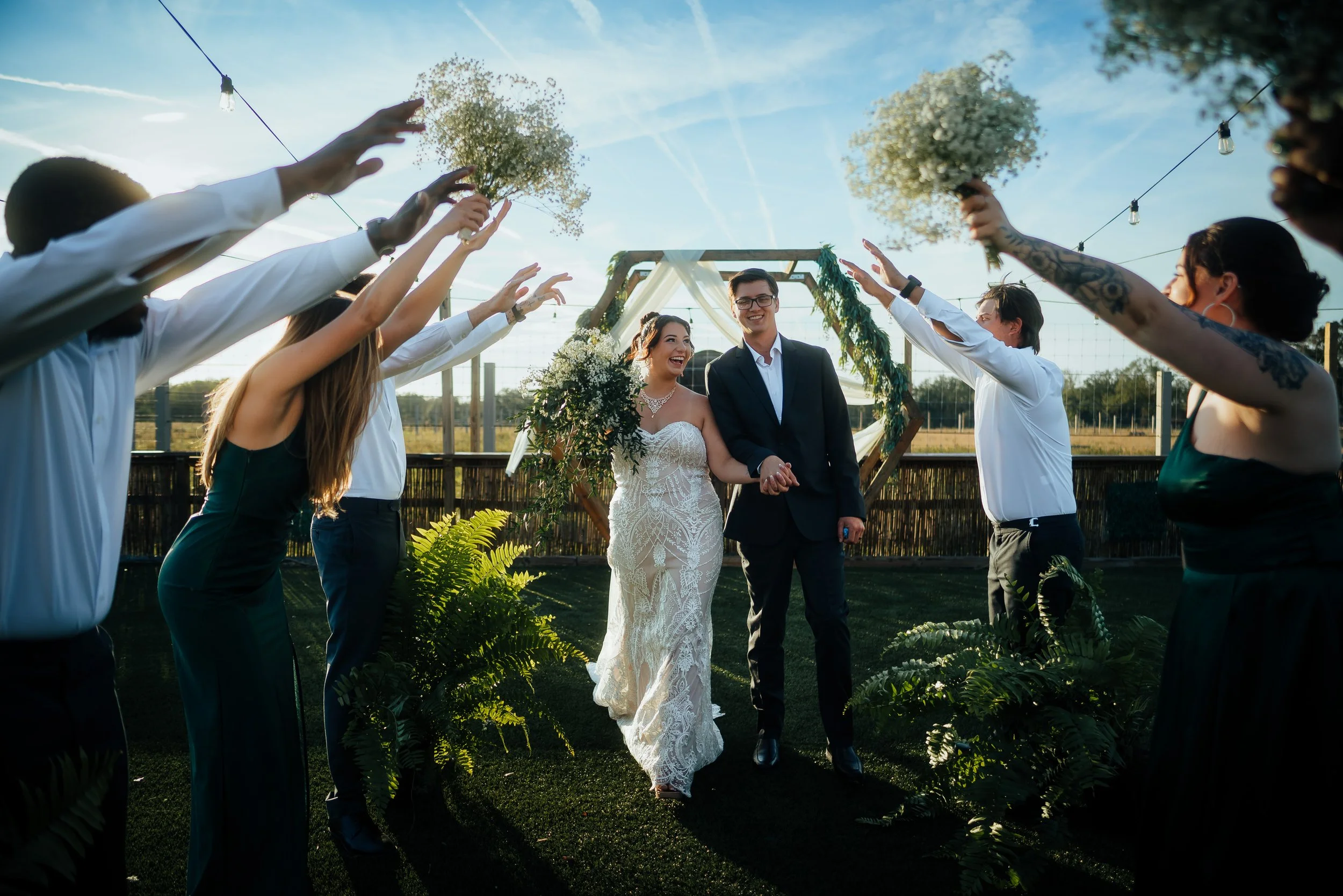 A wedding celebration outdoors at Ivory Acres outside of Sarasota, Florida, with the bride and groom smiling, surrounded by friends holding bouquets of white flowers and reaching towards the couple, under string lights on a sunny day.