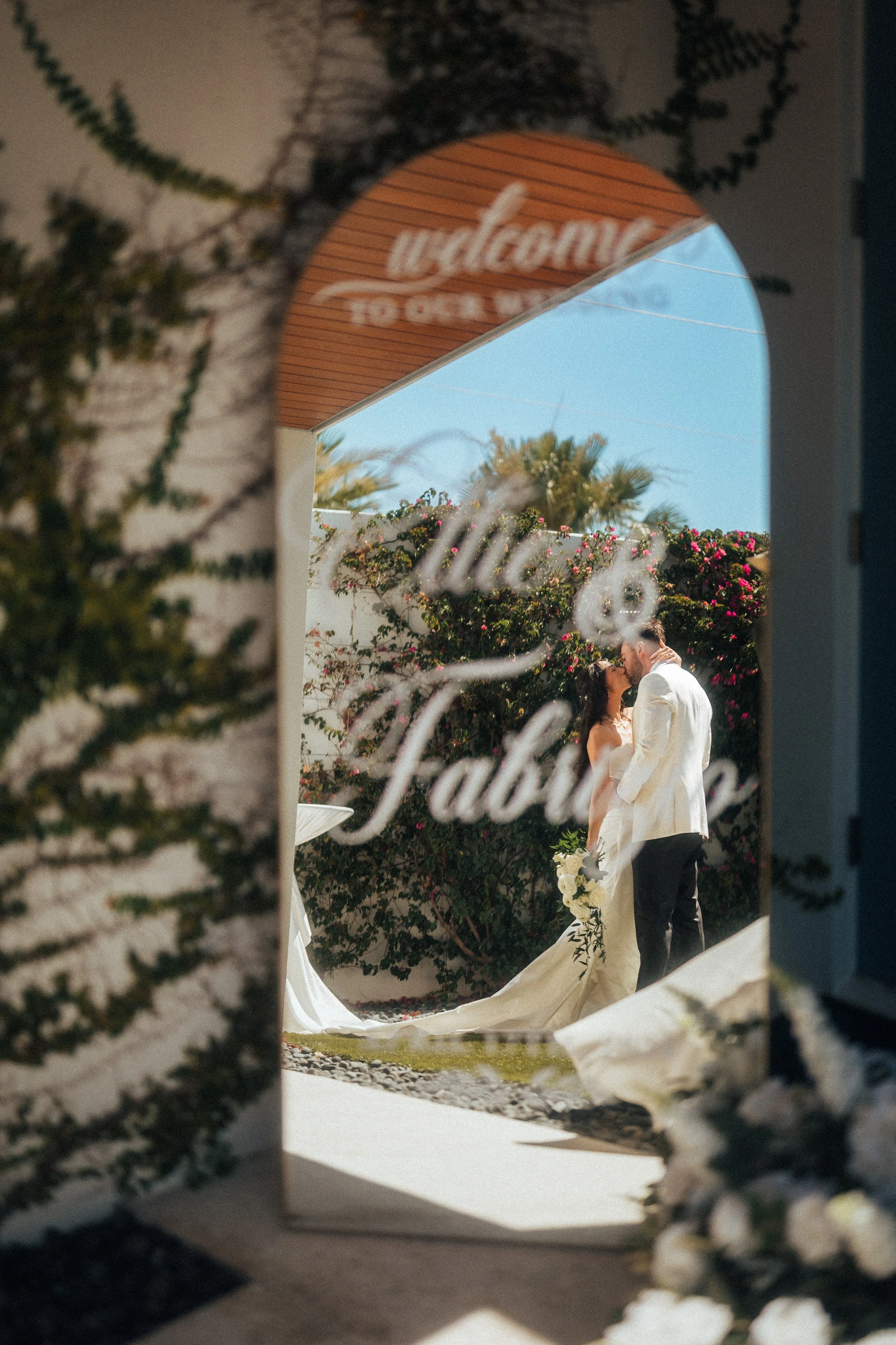 Wedding photo of a bride and groom embrace each other outdoors, seen through a decorative mirror adorned with ivy and overlooking a hedge with pink flowers. It's a bright, sunny Sarasota day.