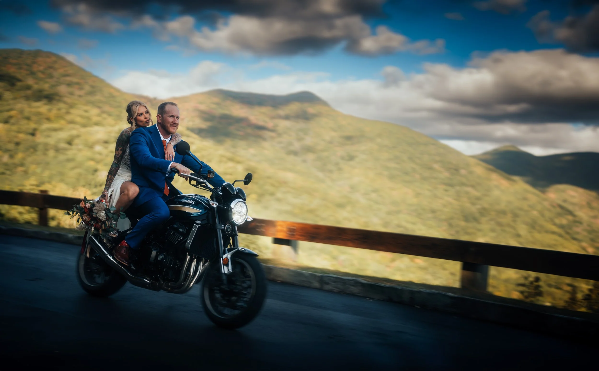 Documentary wedding photo of a newlywed couple riding a motorcycle on the Blue Ridge Parkway with the woman sitting behind the man, holding onto his shoulders, and a backdrop of green mountains and a cloudy sky.