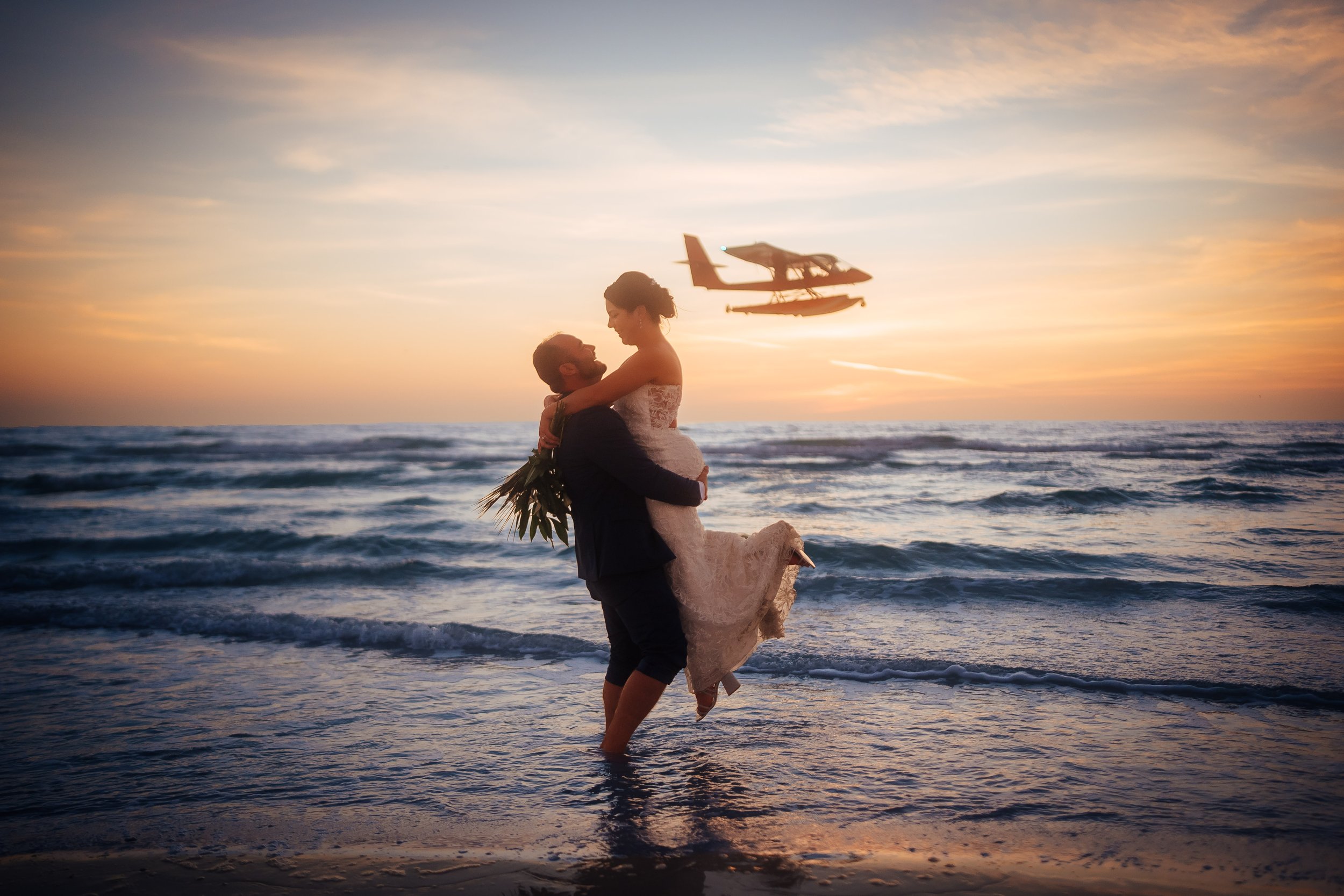 Wedding photo portrait of a couple on Lido Key Beach beach during sunset, with the woman in a wedding dress and the man in a suit, holding each other and smiling. A seaplane is flying in the sky in the background.