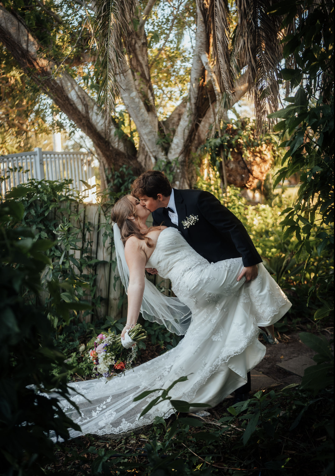 Wedding photo portrait of a bride and groom sharing a kiss outdoors at Palma Sola Botanic Park in Bradenton, with the groom dipping the bride. She is holding a bouquet of colorful flowers, and they are surrounded by green foliage and trees.