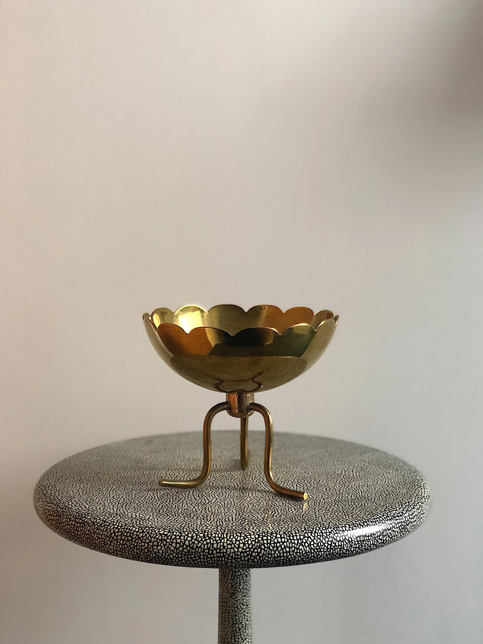 Gold bowl with scalloped edge on a three-legged stand atop a textured round table.