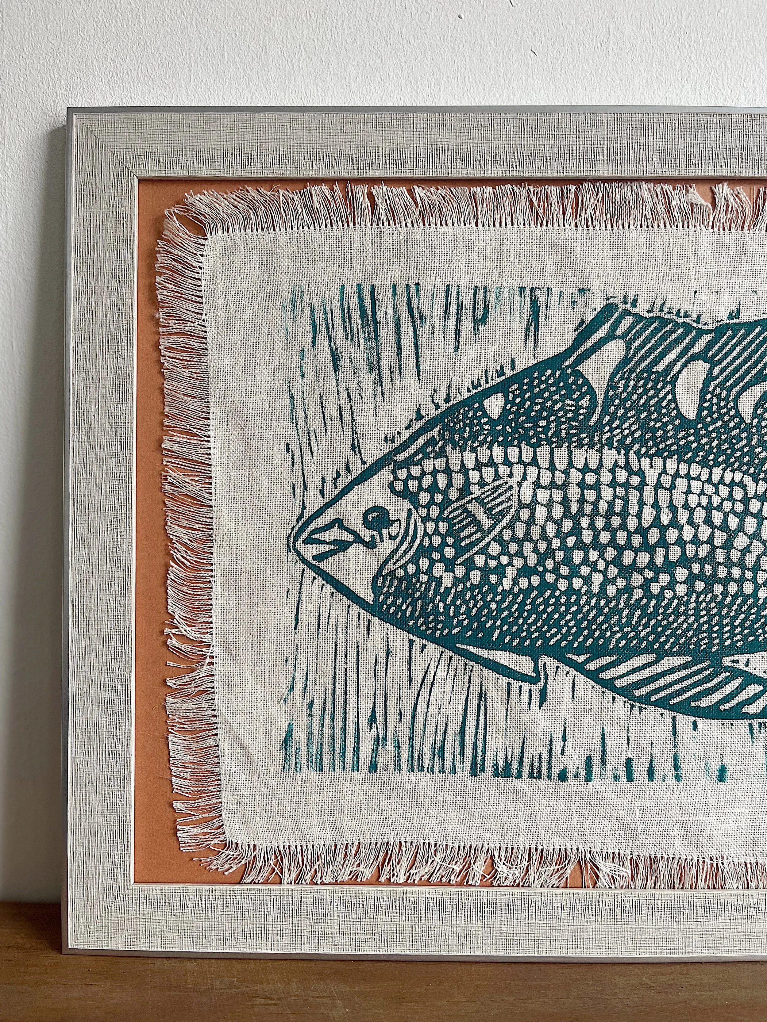 Framed artwork of a stylized fish on textured fabric, with fringed edges.