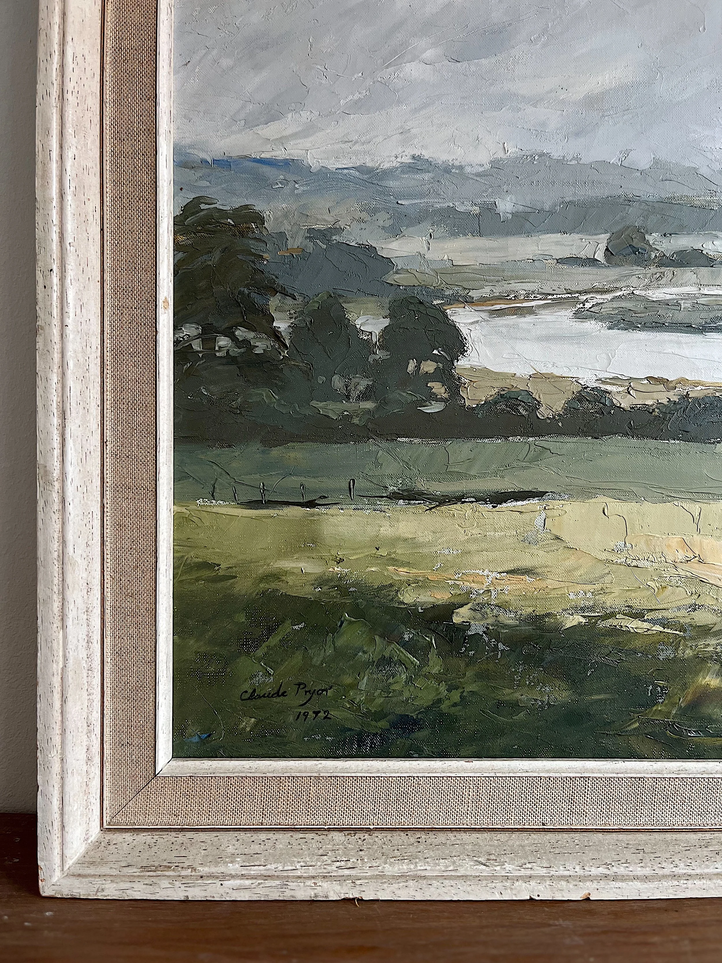 Close-up of a framed landscape painting with a river and trees, signed by Claude Pryor in 1972.