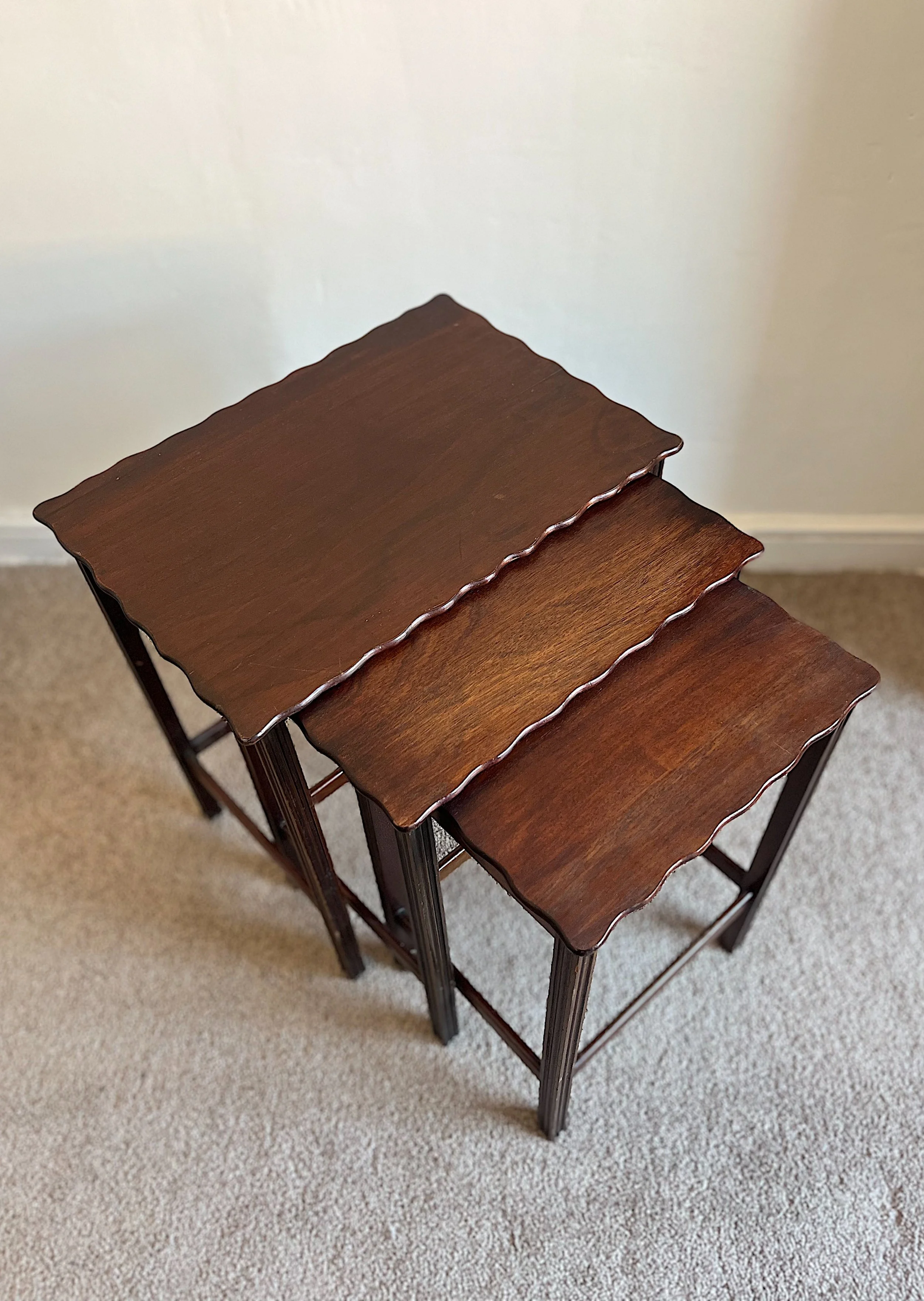 Set of three nesting tables with scalloped edges, made of dark wood, placed on a carpeted floor.