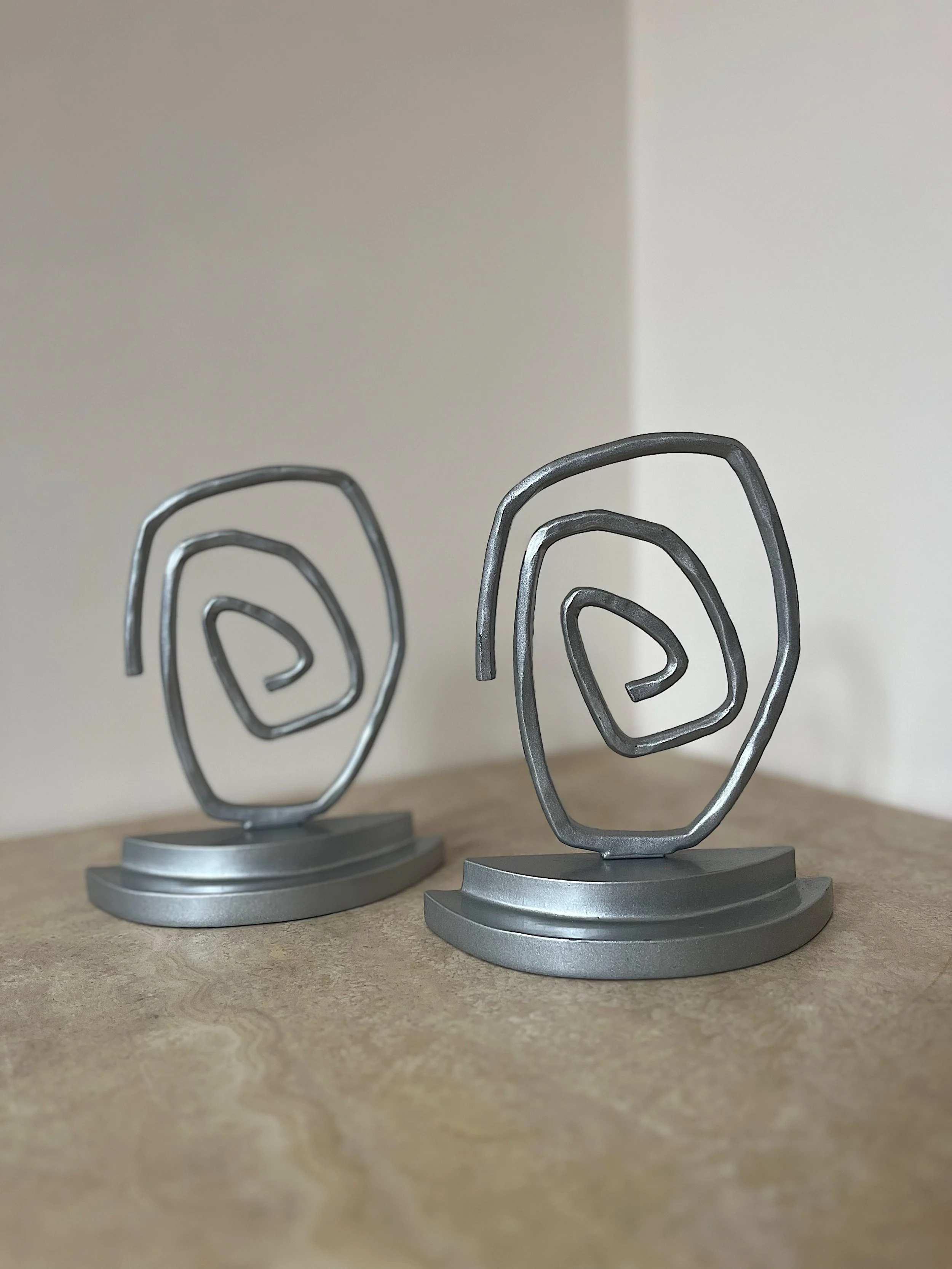 Two abstract metal sculptures on a wooden surface, featuring swirl designs and round bases, set against a neutral wall background.