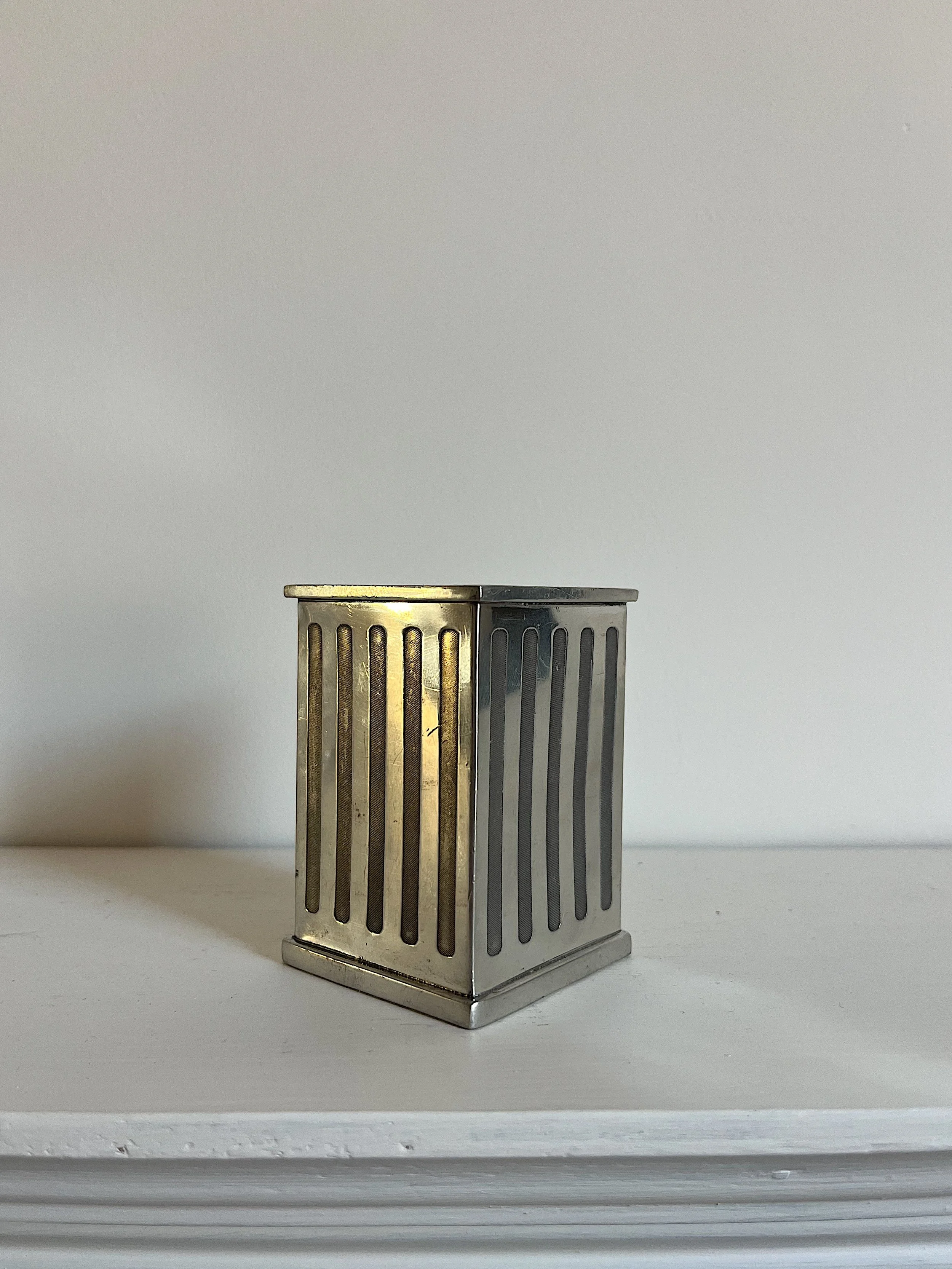 Silver metal decorative container with slotted sides, placed on a white surface against a plain wall.
