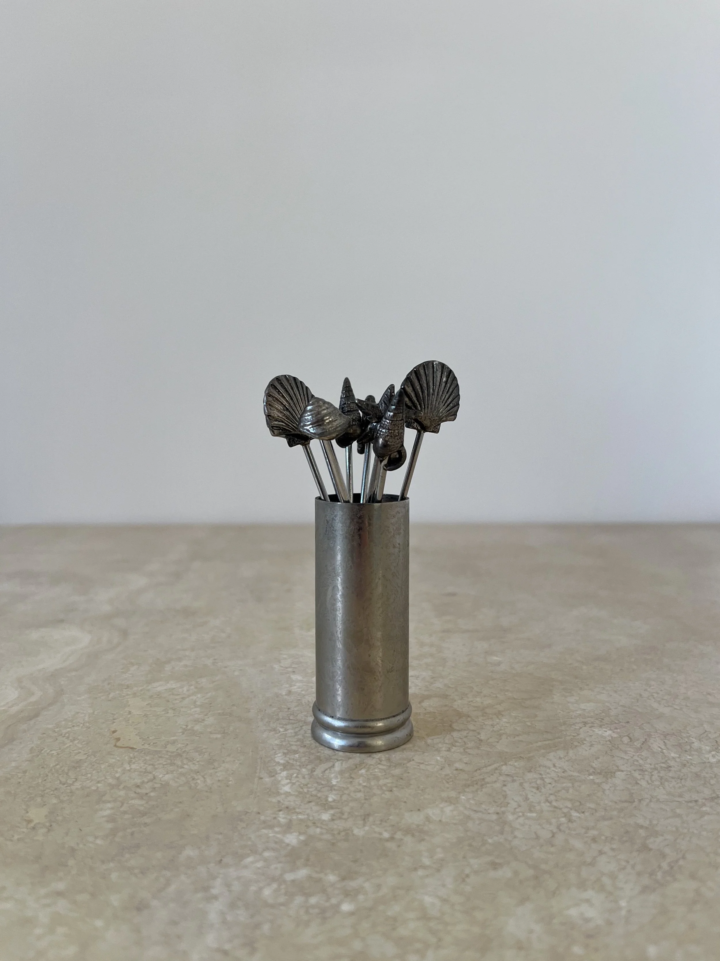 A set of metal cocktail picks with seashell and marine-themed tops, displayed in a cylindrical holder on a light surface.