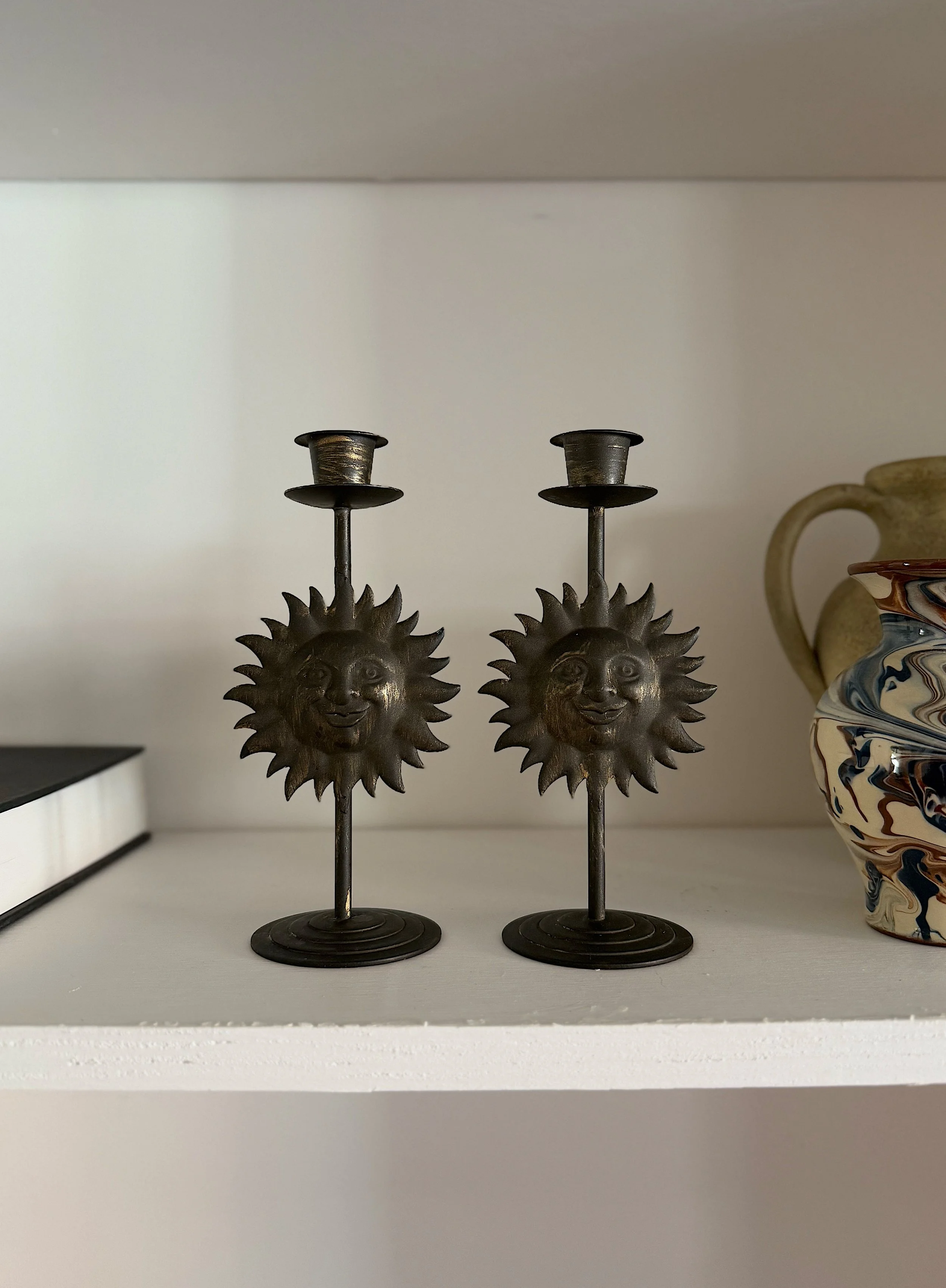 Two sun-shaped metal candlesticks on a shelf between a book and a colorful ceramic vase.