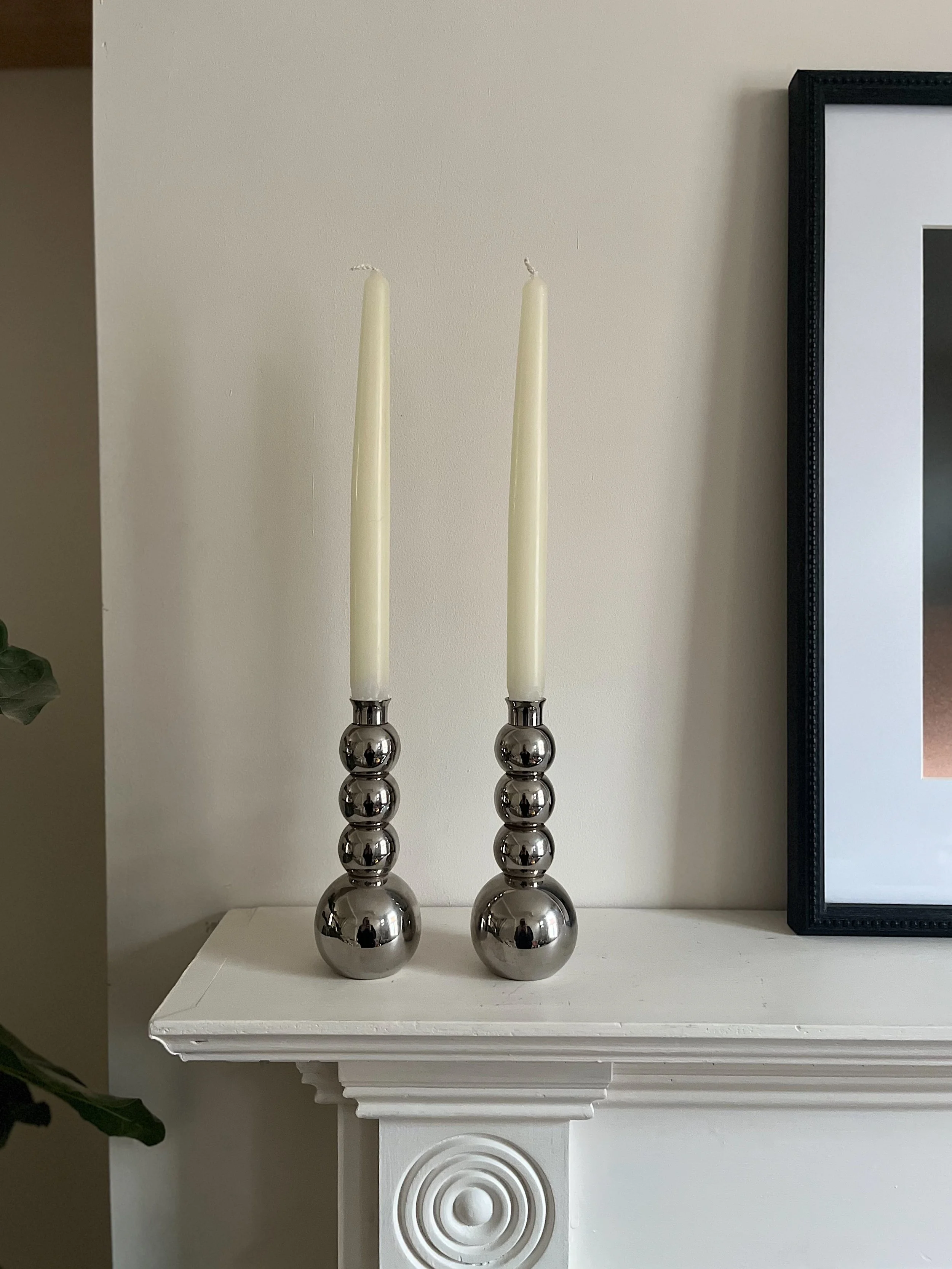 Two silver candlestick holders with white taper candles on a white mantel next to a framed picture.