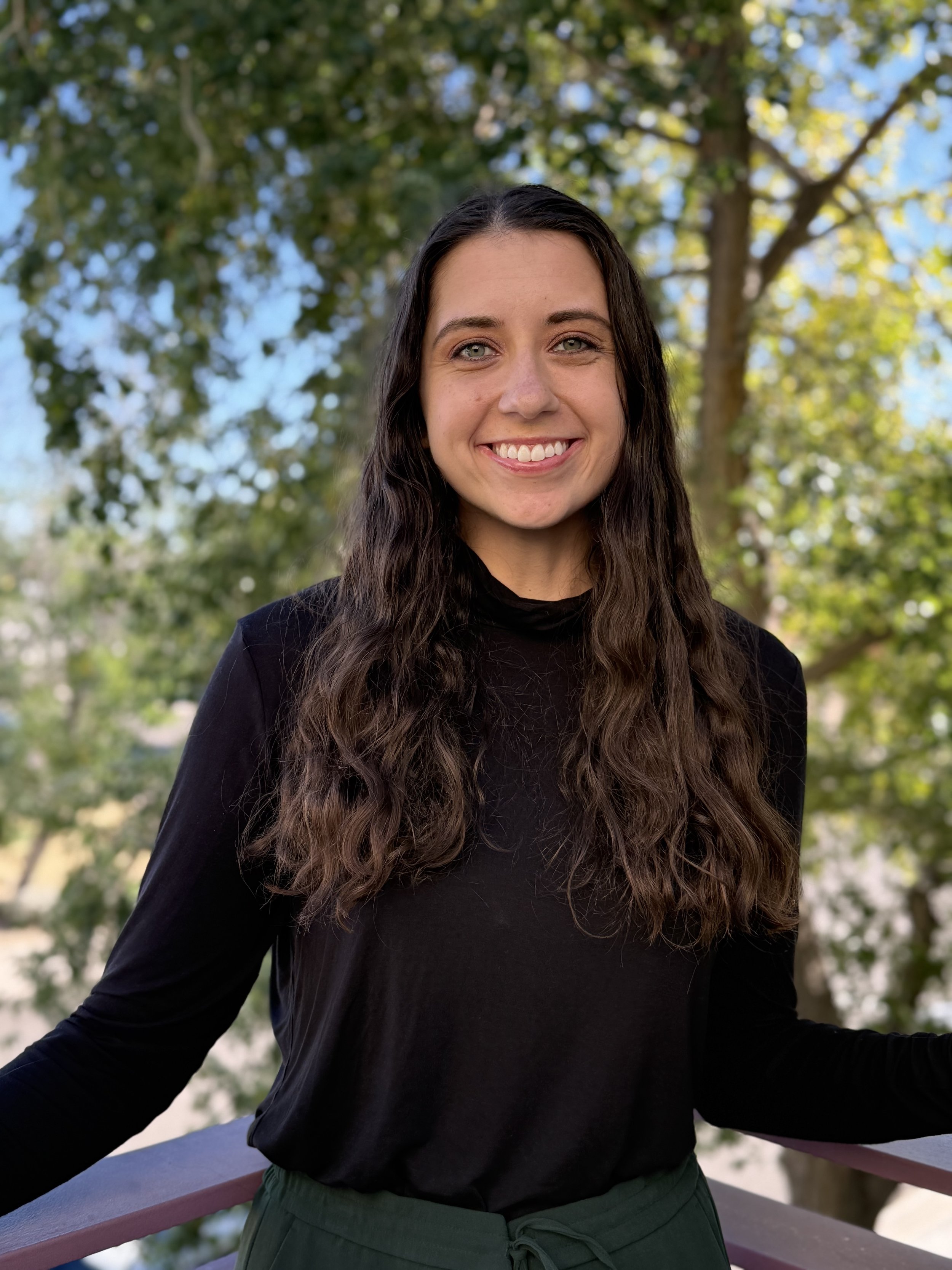 A young woman with long wavy brown hair, smiling, standing outdoors on a sunny day with green trees in the background, wearing a black long-sleeve top and green pants.