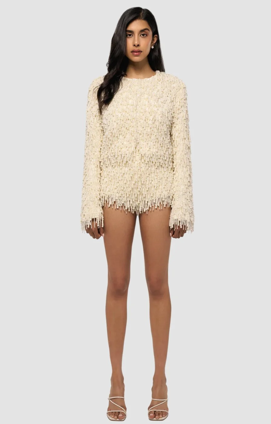 A woman with long dark hair wearing a cream-colored, textured, fringe-detail sweater, standing against a plain gray background.