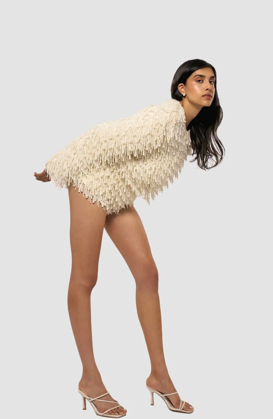 A woman with long dark hair and a serious expression is wearing a cream-colored textured sweater and beige high-heeled sandals, standing against a plain light gray background.