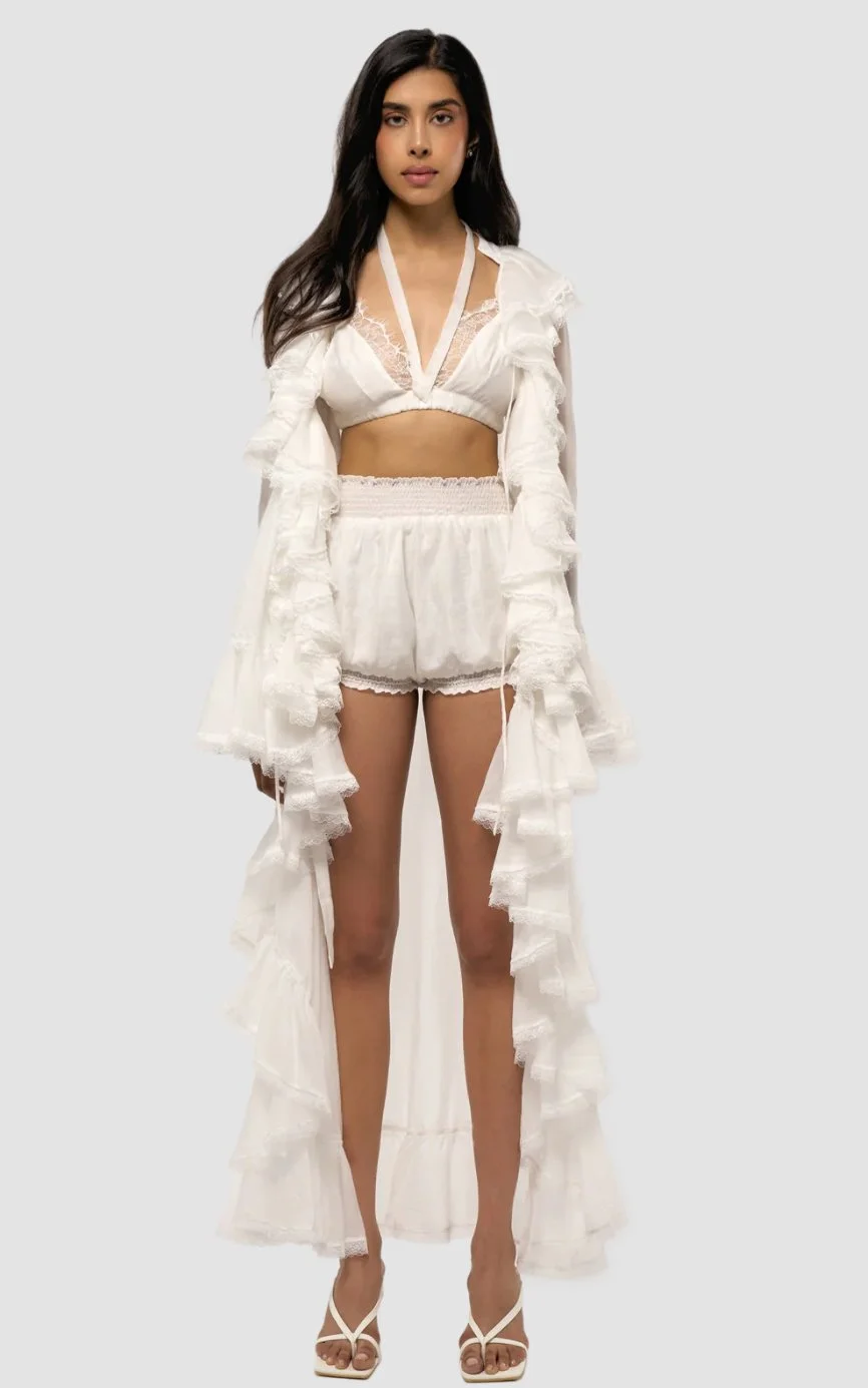 A woman in a white bridal or fashion ensemble, featuring a crop top, shorts, and a long ruffled cape, standing against a plain background.