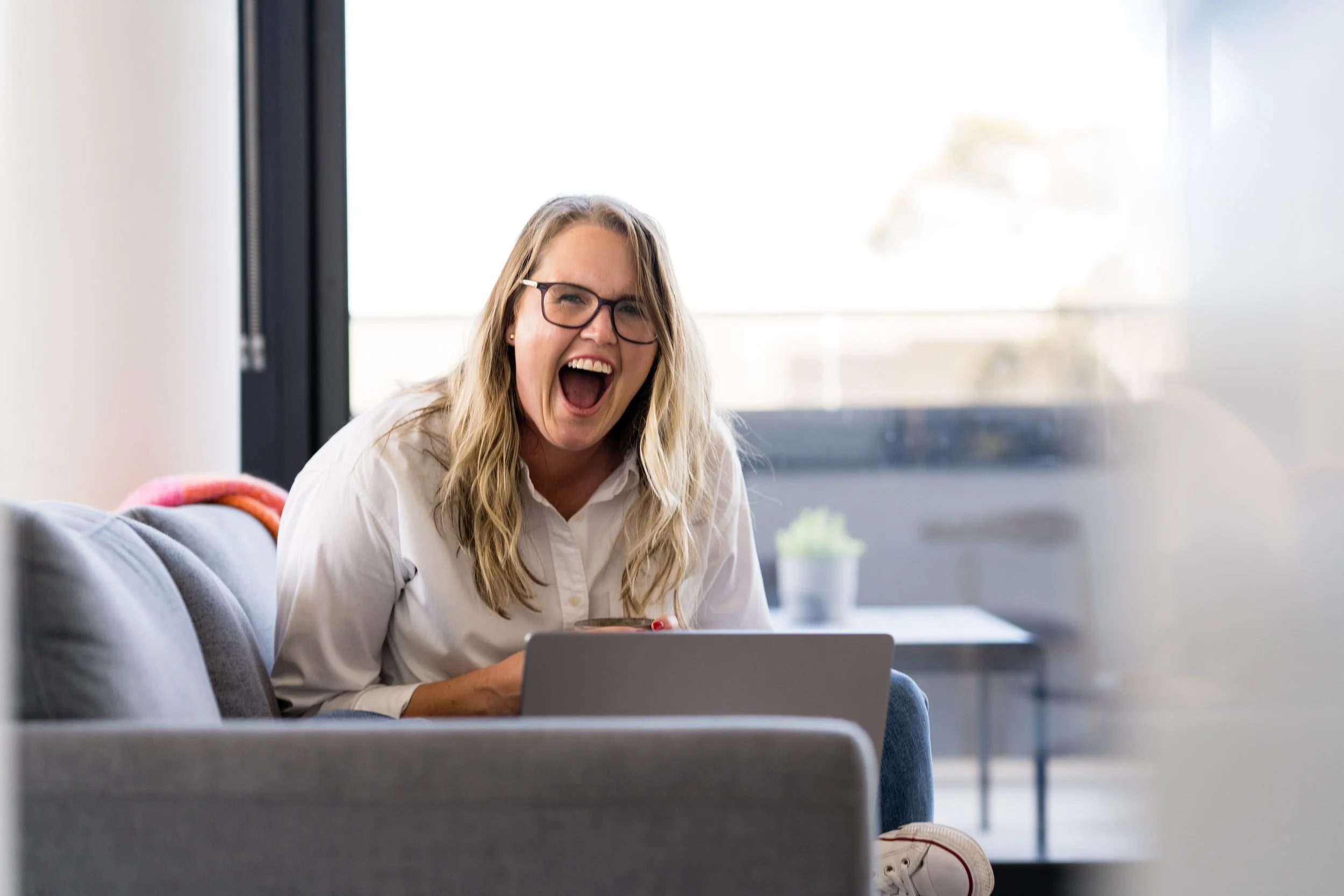 Kathy Rast sits on a grey sofa and is laughing to large we swear you can see her larynx