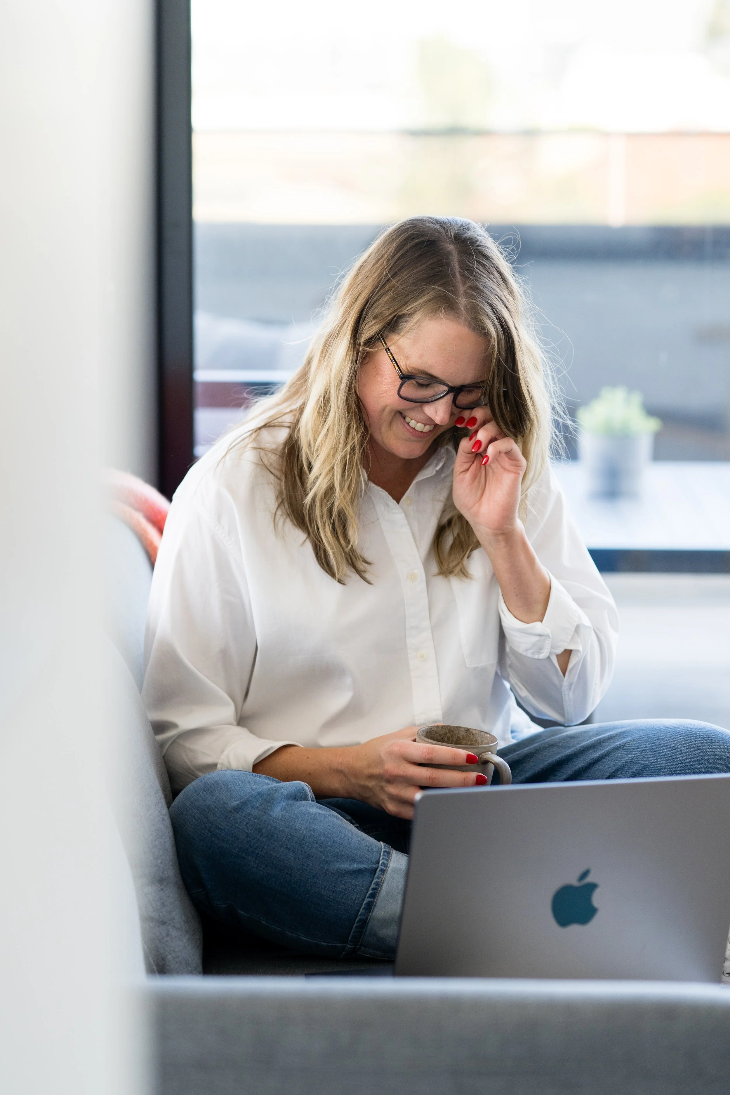 Kathy Rast holds a mug she in one hand and adjusts her glasses with the other as she sits on a grey sofa working on her laptop