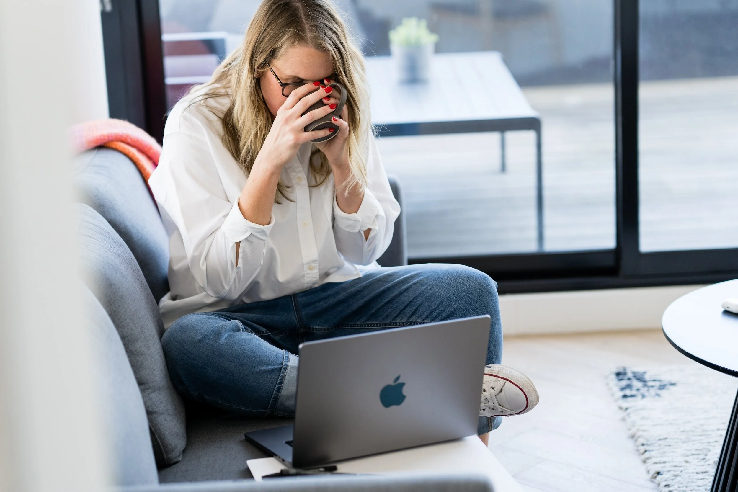 Kathy Rast drinks from a mug she holds in both hands while sitting on a grey sofa working on her laptop