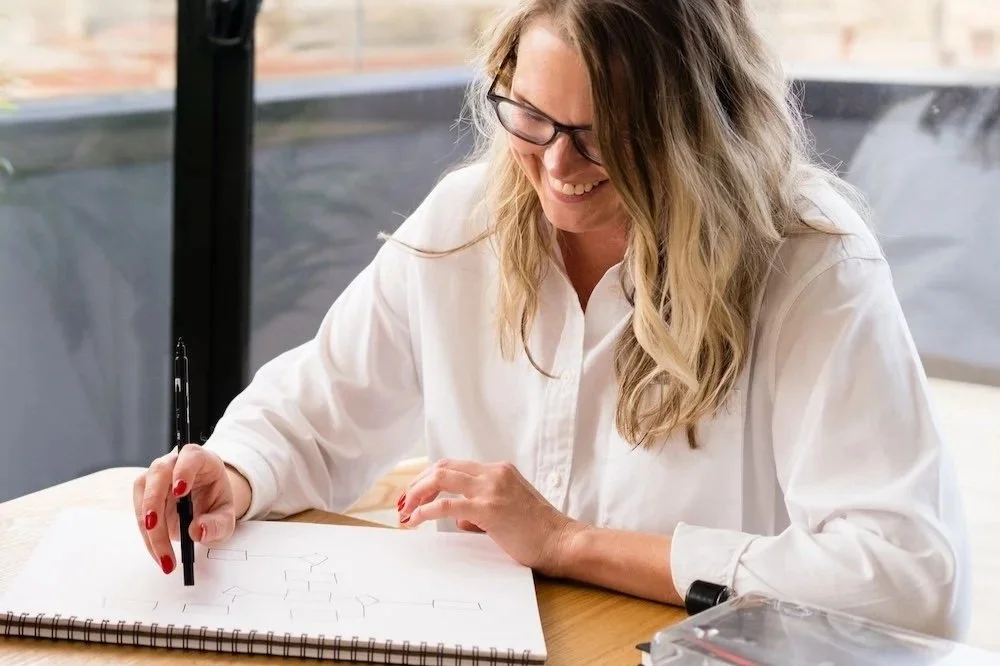 Image - Kathy Rast sits at a wooden table with a large notebook and black marker as she maps her thinking onto a white page
