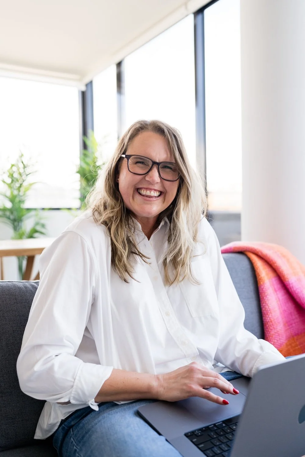 Kathy Rast is sitting on grey sofa with her laptop resting in her lap all while she smiles the weirdest smile you can image - it's weird and kinda wonderful