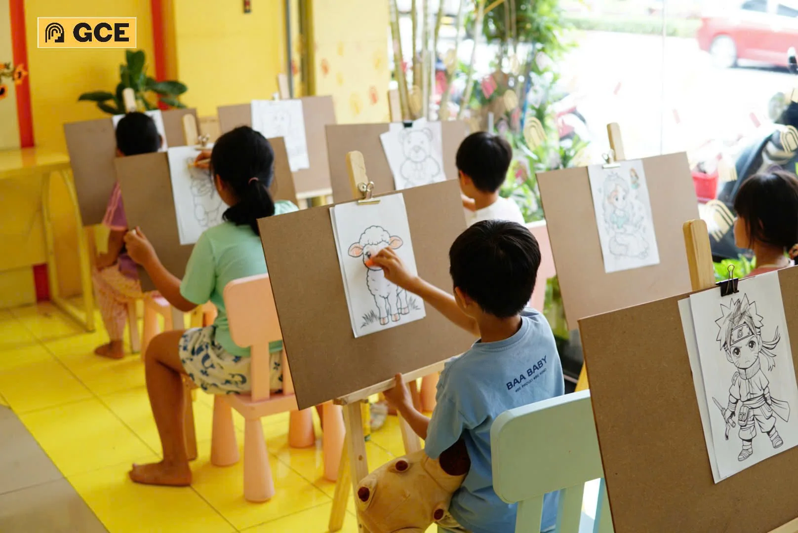 Children sitting at easels coloring pictures of cartoon characters and animals in a classroom with yellow walls and large window.