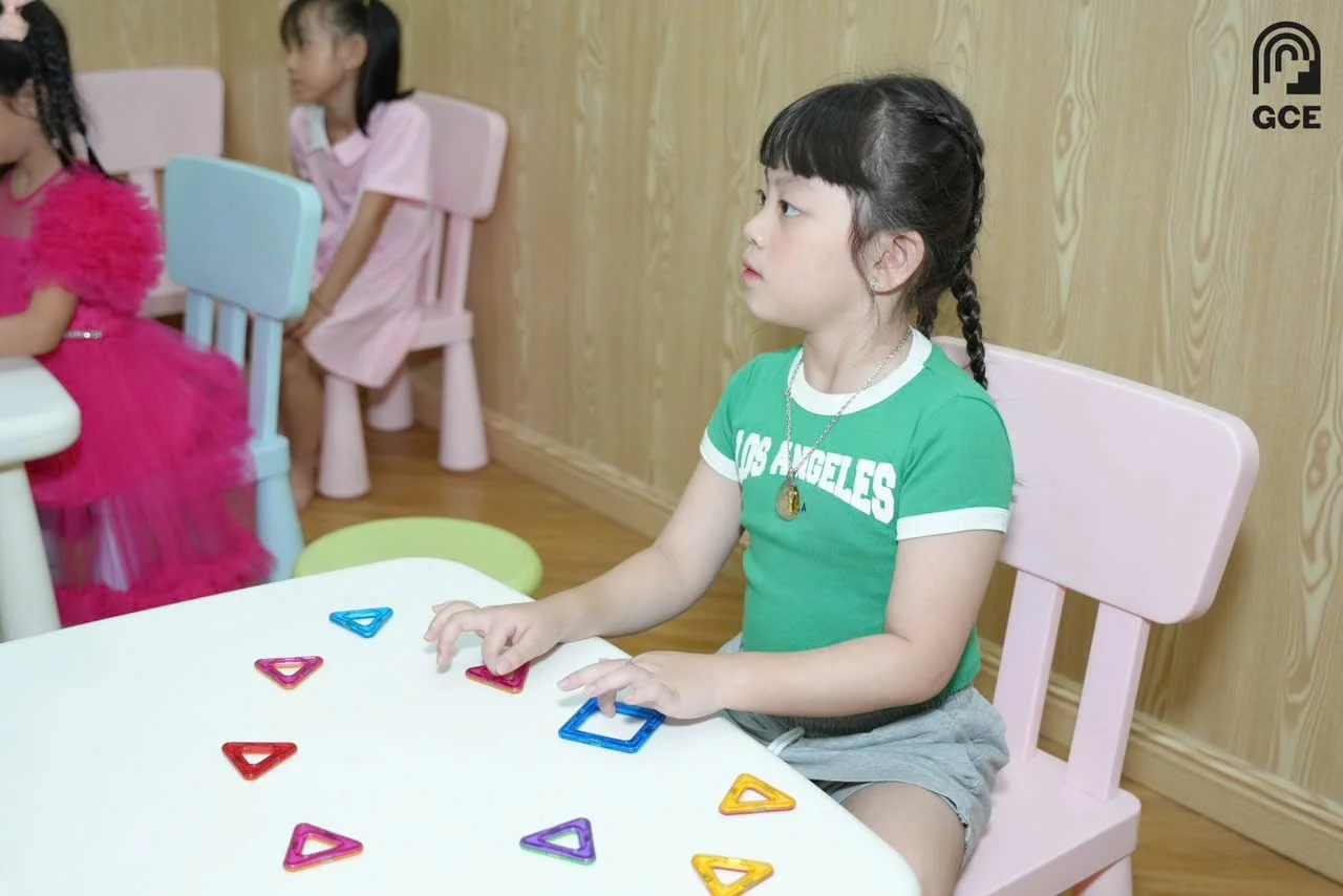 A young girl wearing a green 'Los Angeles' t-shirt and gray shorts is sitting at a white table with colorful triangular and square magnetic tiles, in a room with a wooden wall and pink chairs, while other children are seated nearby.