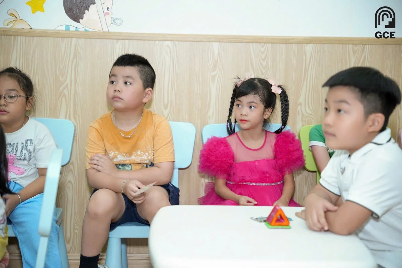 Group of children sitting at a table in a classroom, with a boy in an orange shirt, a girl in a pink dress, and other children in the background.