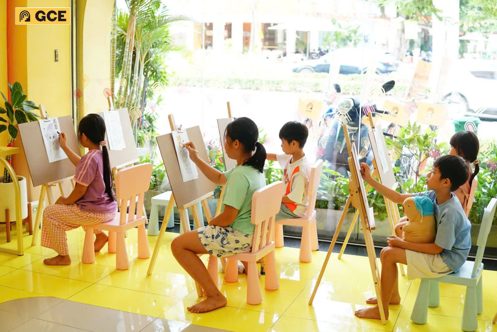 Children sitting on chairs, drawing on easels, in a brightly lit art classroom near a large window with plants outside.