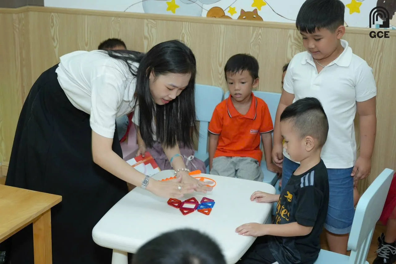Teacher demonstrating something to four children gathered around a table with colorful geometric shapes in a classroom.