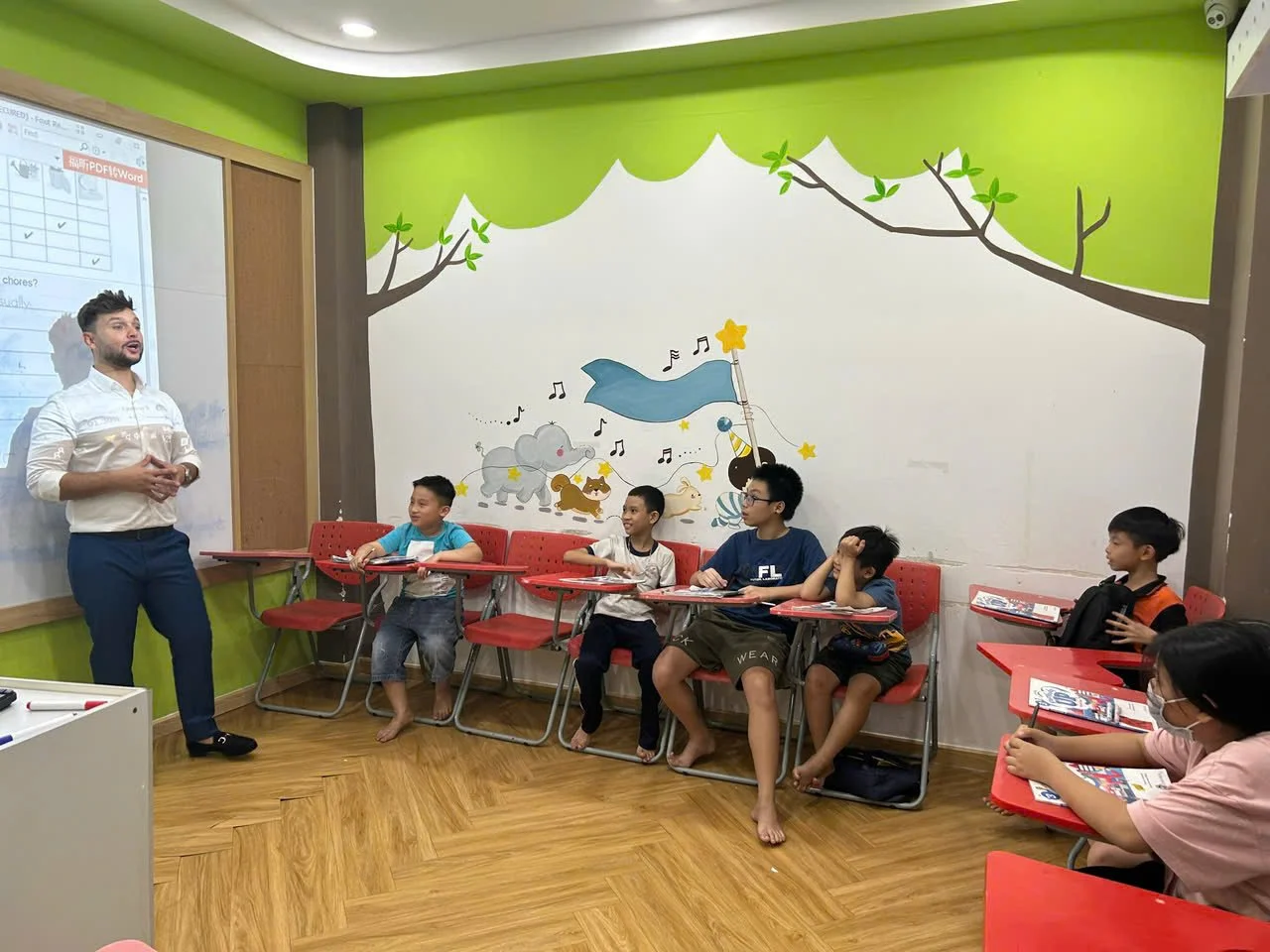 A teacher standing in front of a classroom of children sitting at red desks with school supplies, a teacher explaining something to students, a colorful wall mural of animals and music notes behind them.